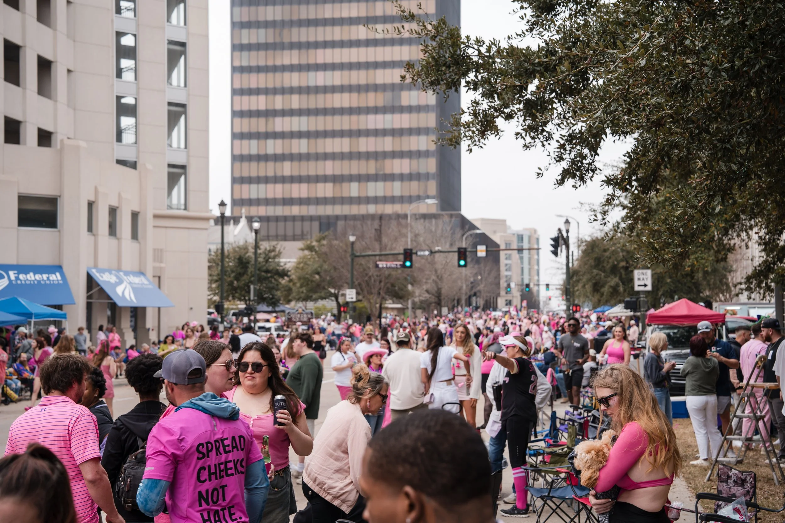 A crowd of people at a street event, many dressed in pink, with some holding drinks and talking. There are tents and high-rise buildings in the background, and a traffic light overhead.