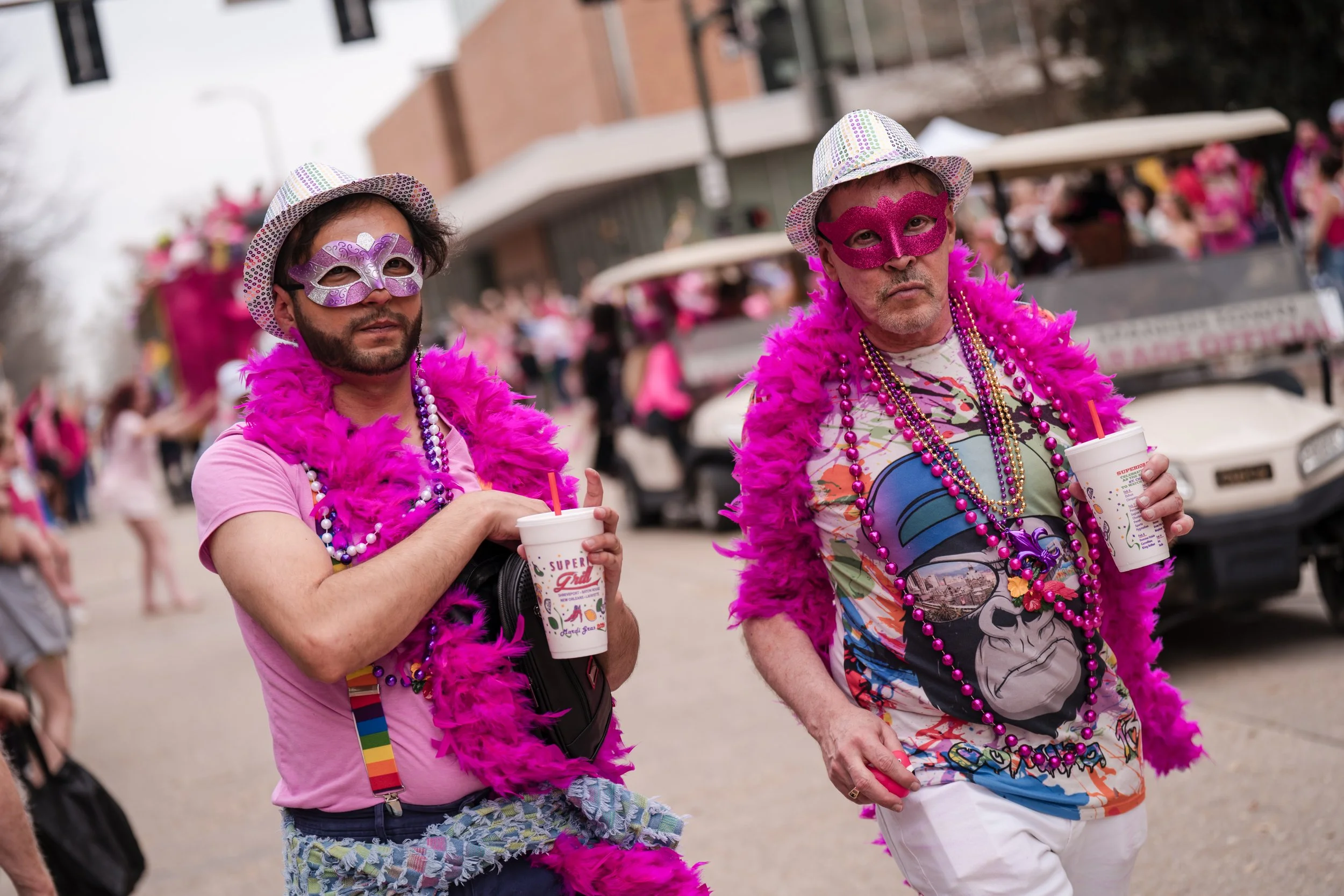 Two men dressed in colorful outfits with pink feather boas and masks, holding large drinks with straws, participating in a parade or festival celebrating Mardi Gras or a similar festive event.