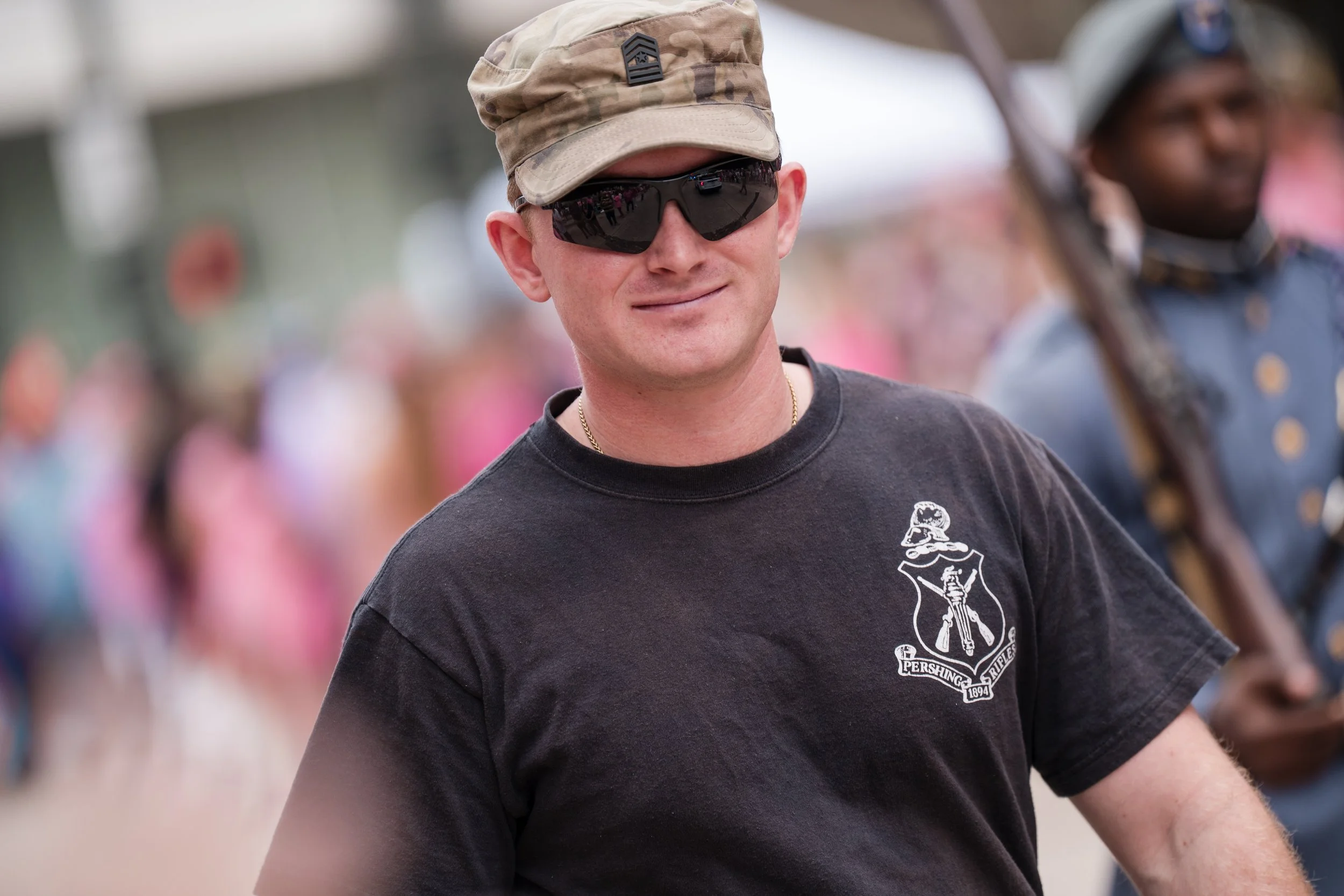 A smiling man wearing sunglasses, a camouflage cap, and a black T-shirt with a crest emblem on the chest, standing outdoors at a crowded event.