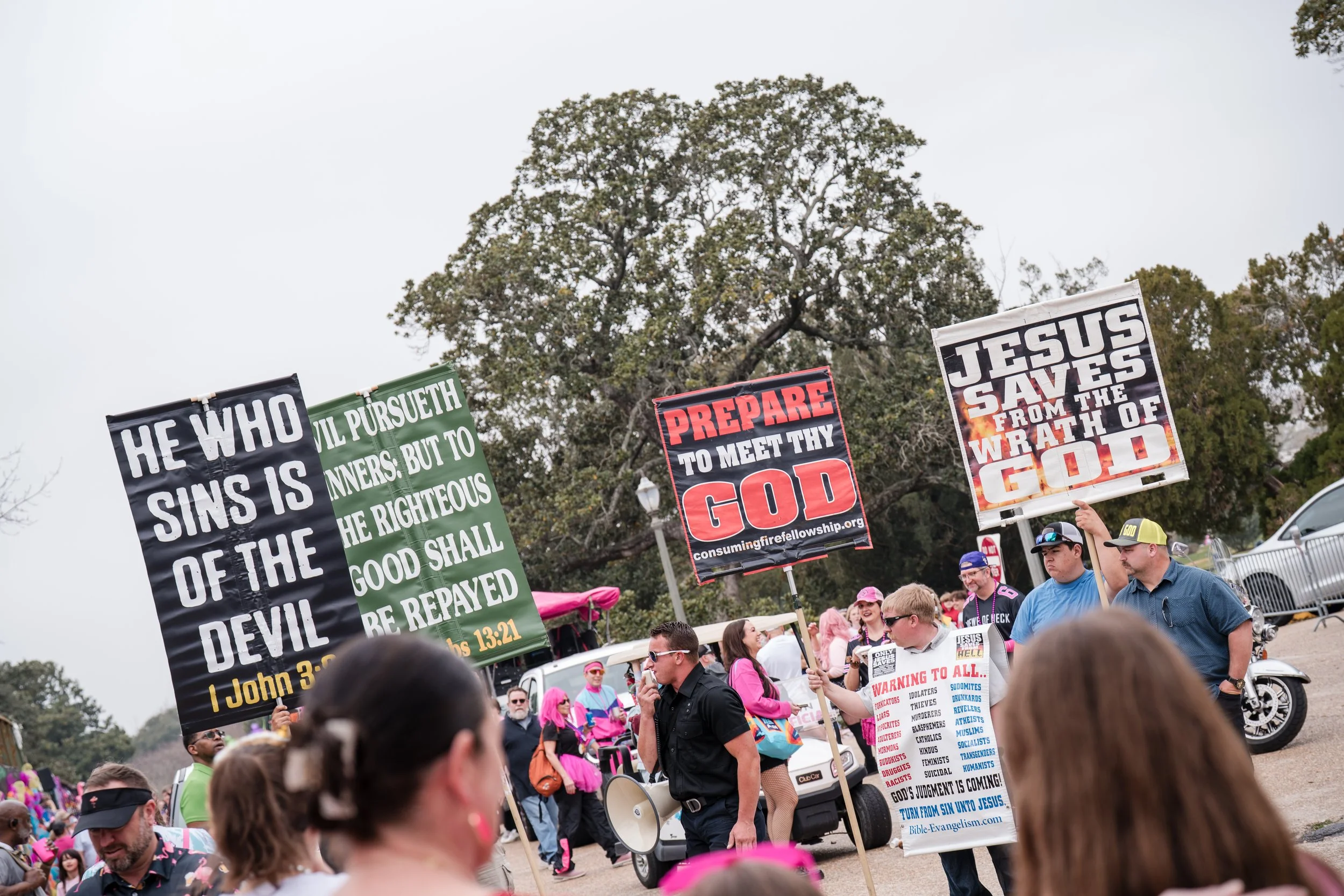 People participating in a protest or rally holding signs with religious messages about Jesus, God, and sin, with large trees and a parking lot in the background.