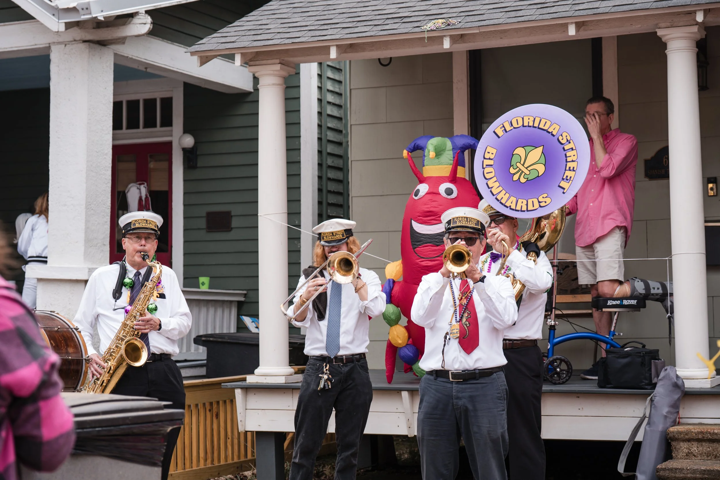 A band of musicians wearing Florida Street Bowhards hats is playing instruments on a porch during a festive event. Behind them, a person in a pink shirt stands beside a colorful giant inflatable caterpillar.