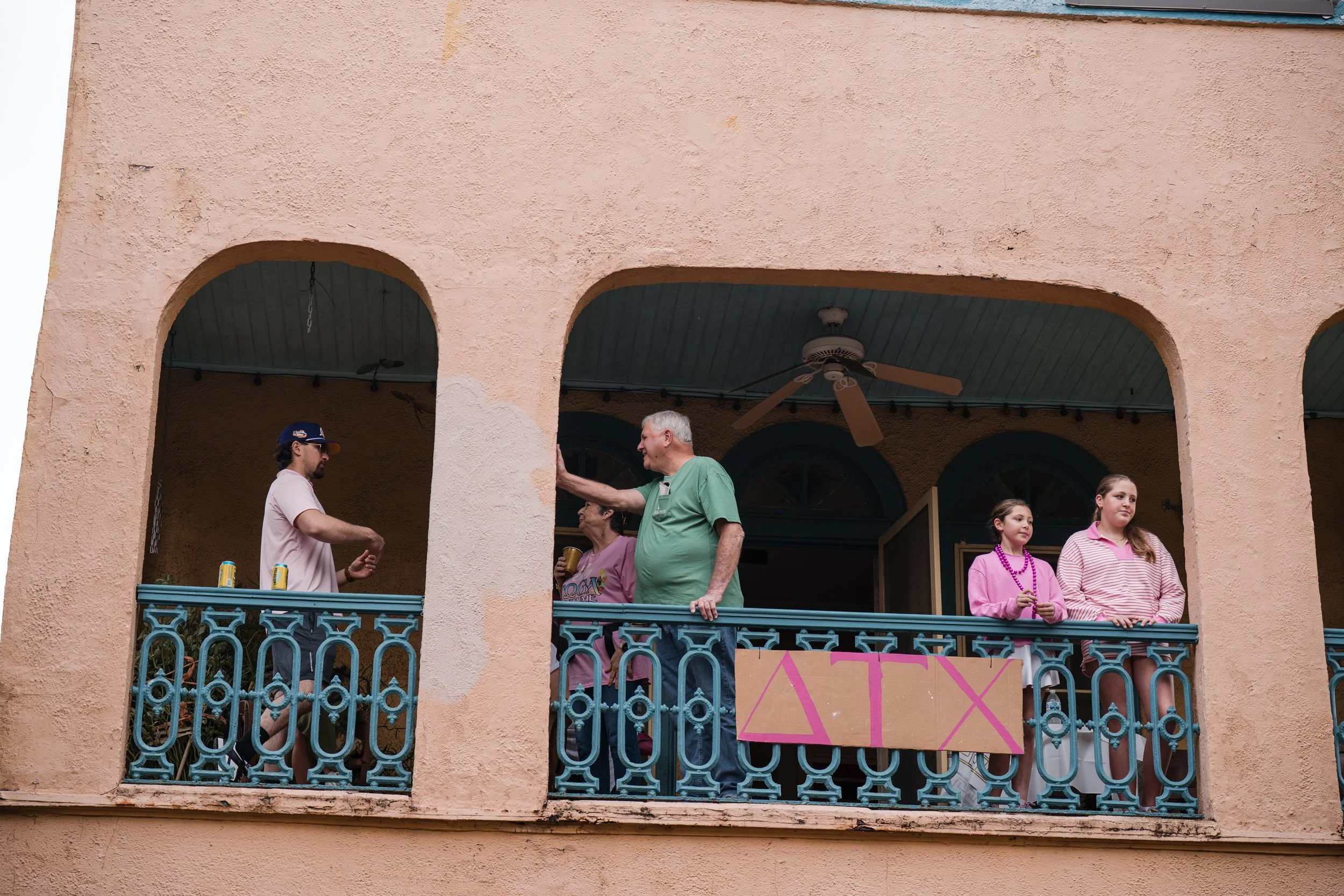 Five people are standing on a balcony with a pink wall and blue ornate railing. Two young women are on the right, one in a pink striped shirt and the other in a purple shirt. Two men are in the middle, one older man touching the wall and a younger ma