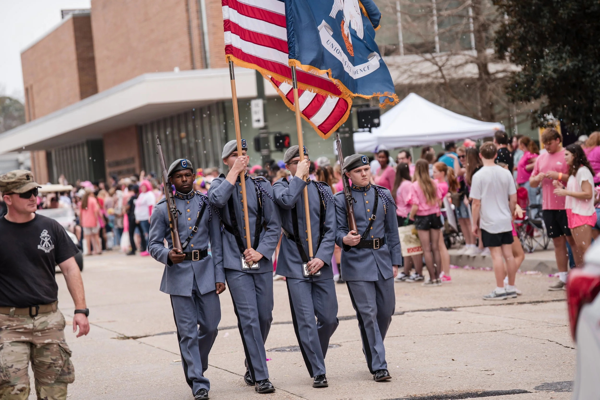 Four uniformed military personnel marching in a parade, carrying an American flag and a state flag, with spectators and pink-shirt participants in the background.