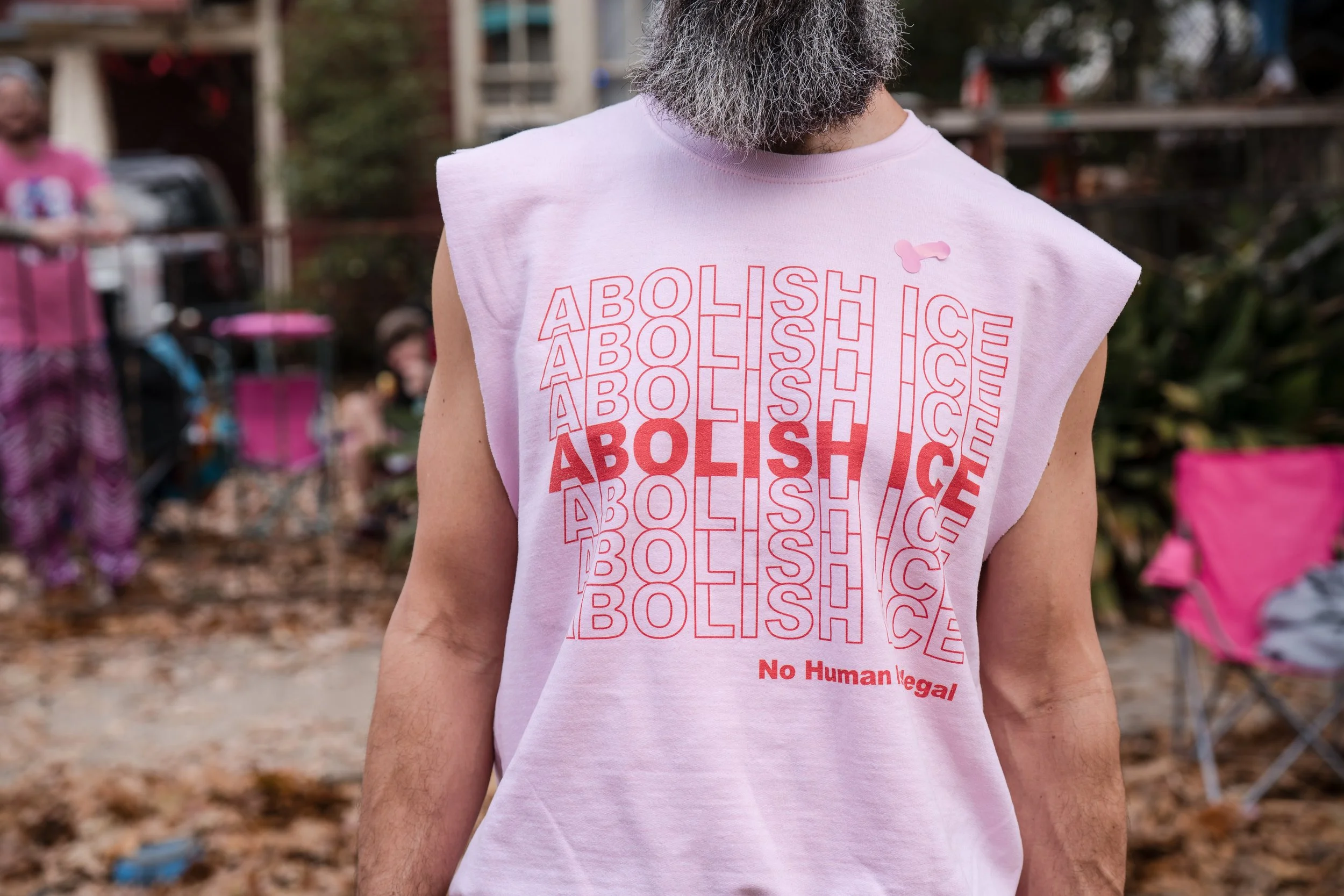 Man wearing a sleeveless pink T-shirt with the words 'ABOLISH ICE' repeated in red text, with the phrase 'No Human Illegal' printed at the bottom. The man has a gray beard, and there are people and trees in the background.