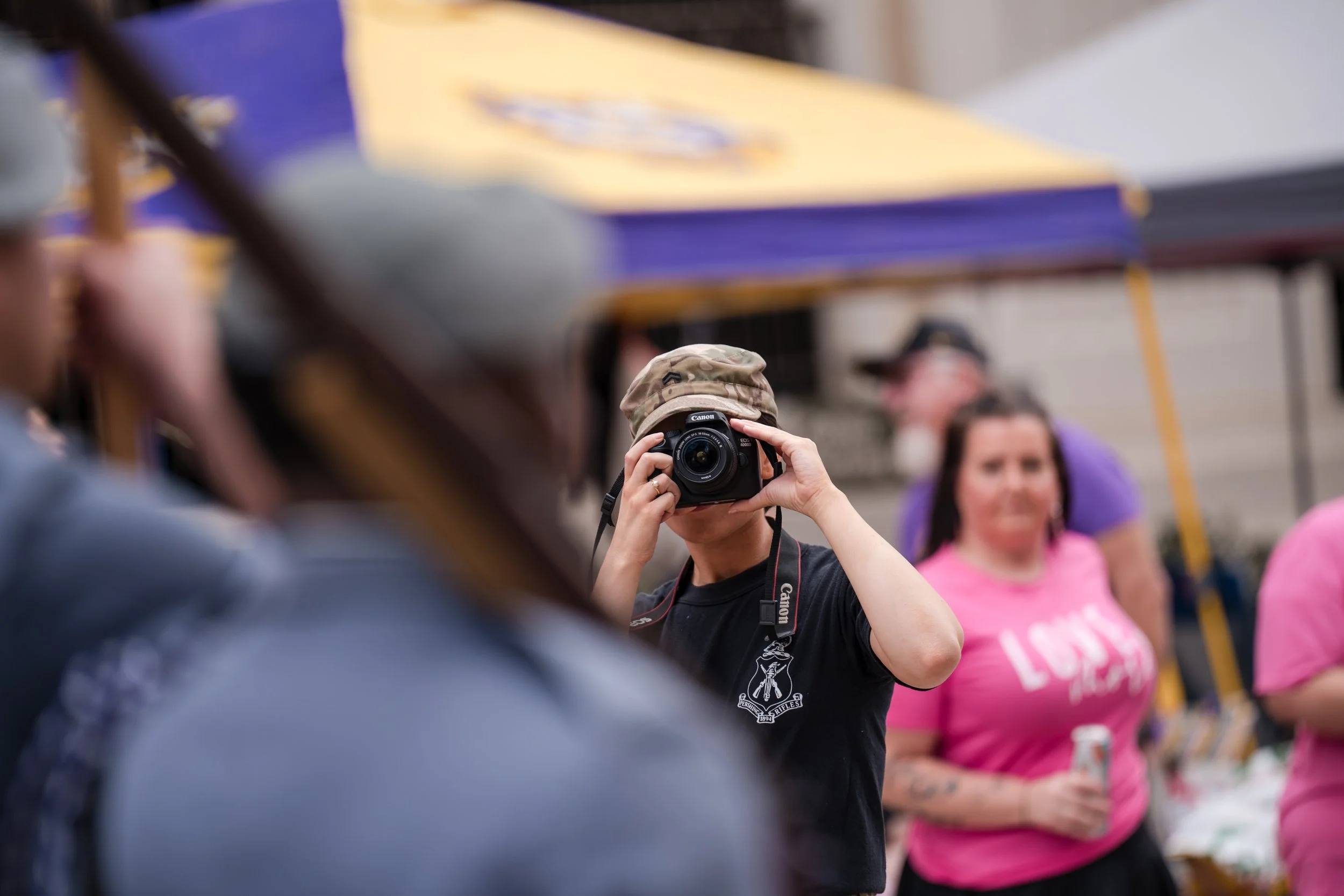 Person wearing camouflage hat taking a photo with a camera at an outdoor event, with several blurred people and tents in the background.