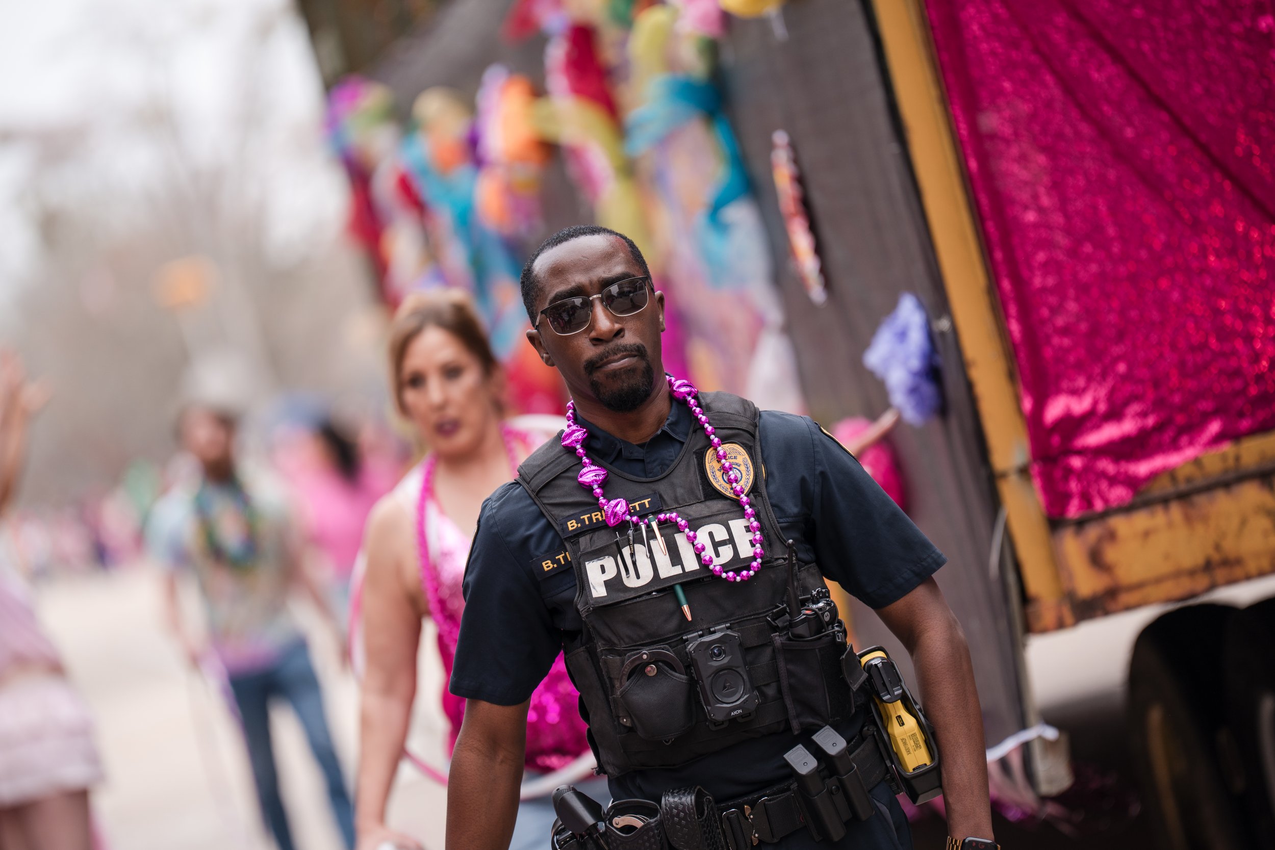 A police officer in uniform wearing sunglasses and a purple bead necklace, standing on a street during a parade or celebration, with people and colorful floats in the background.