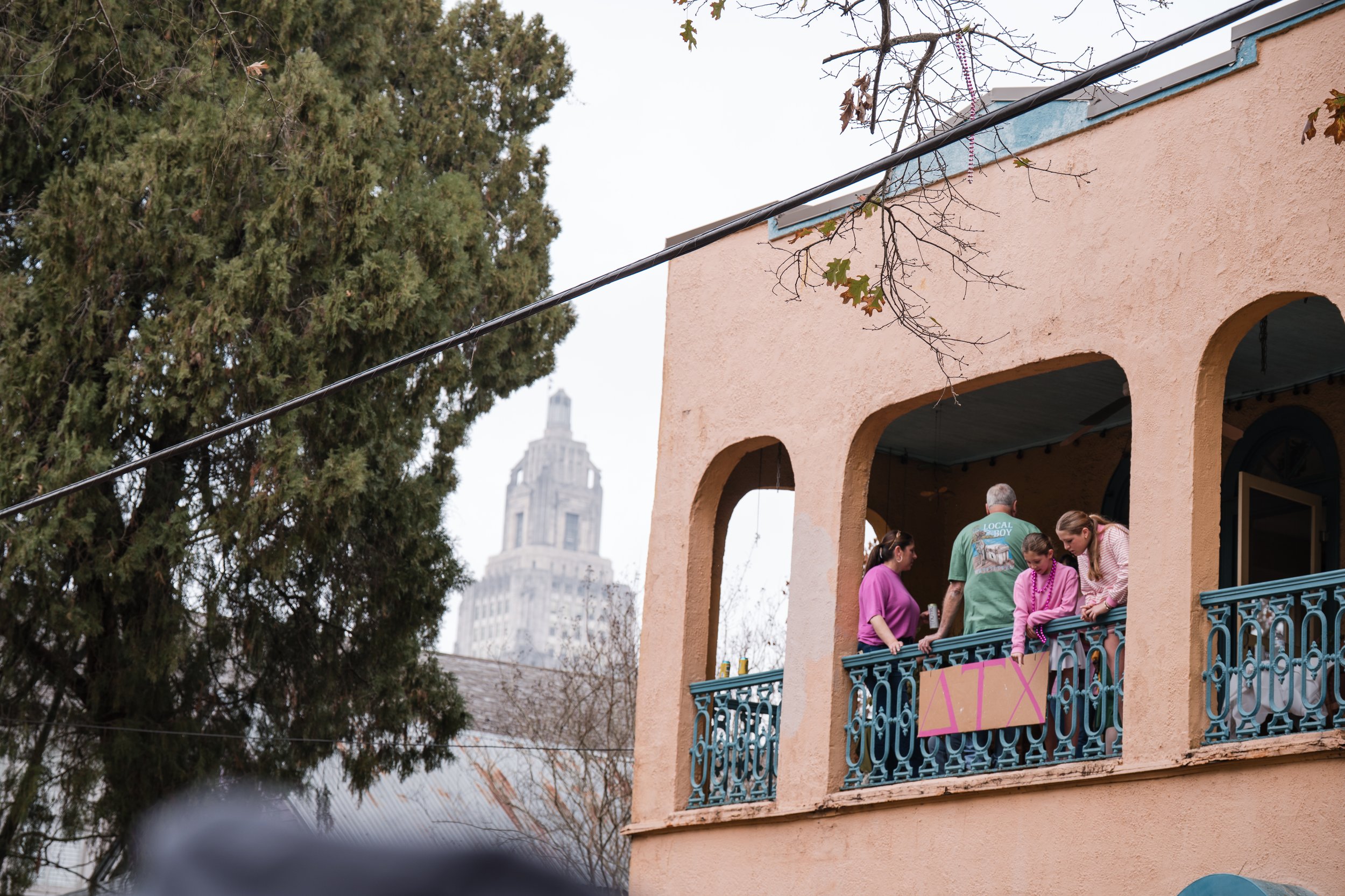 People gather on a peach-colored balcony of a building with arched openings, overlooking a city height with a tall historic building in the background.