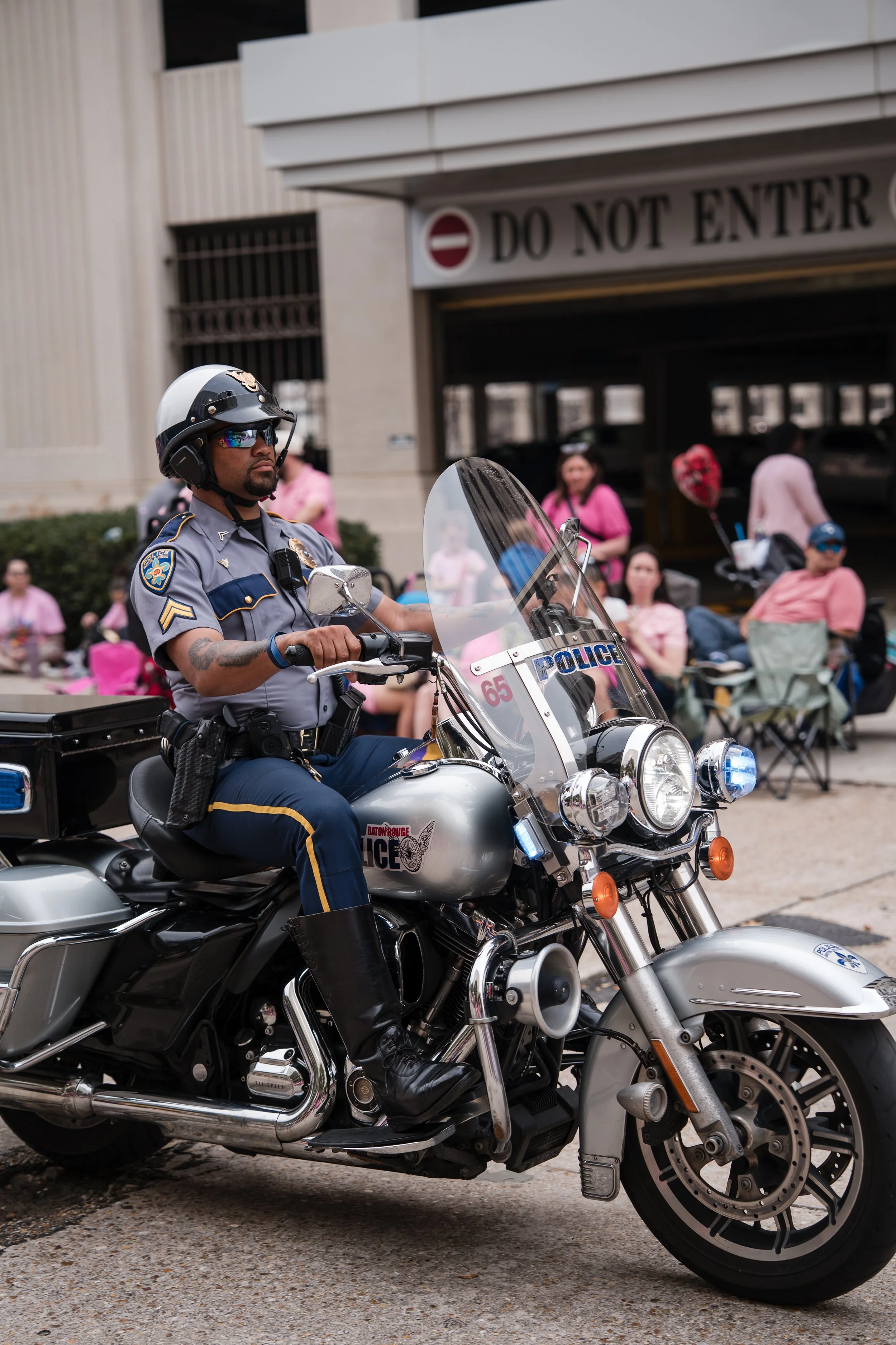 A police officer riding a motorcycle during a public event, with people sitting and standing in the background.