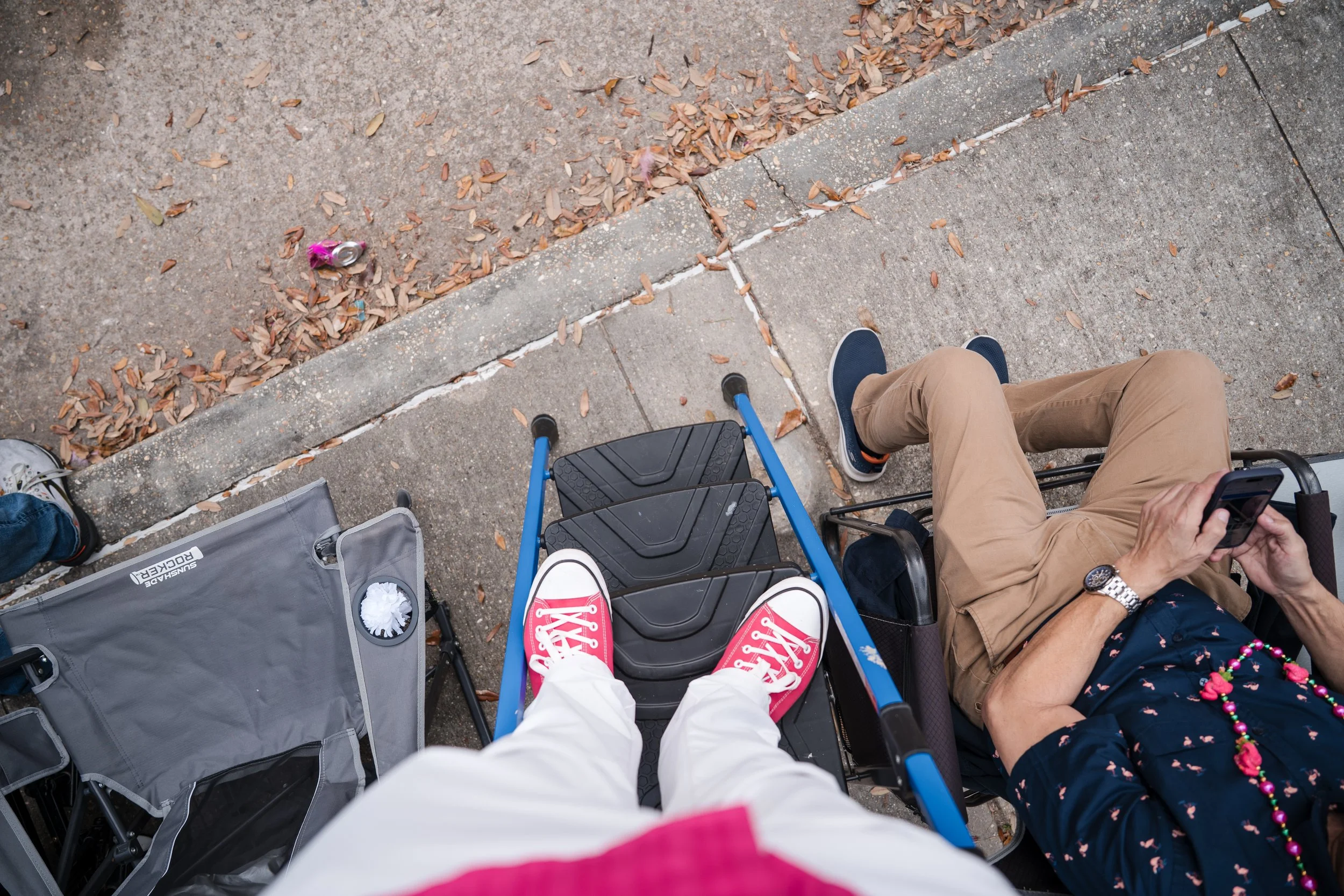 Overhead view of two people sitting in wheelchairs next to each other on a sidewalk; one person wearing red sneakers and second person using a phone; fallen leaves and trash on the ground.