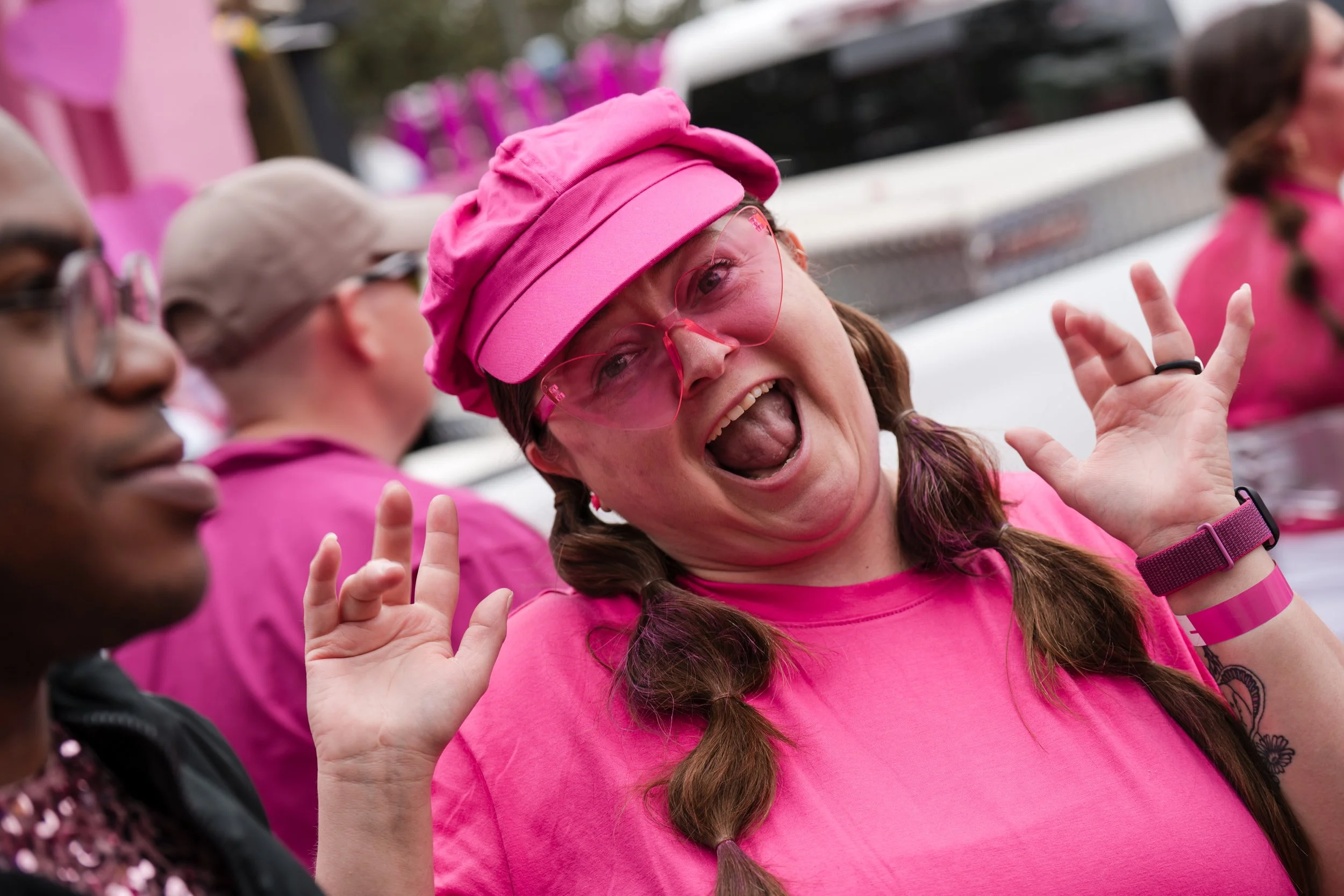 A woman wearing a pink cap, pink-tinted glasses, and a pink shirt, laughing and making a playful gesture with her hands at an outdoor event. Other women in pink are visible in the background.