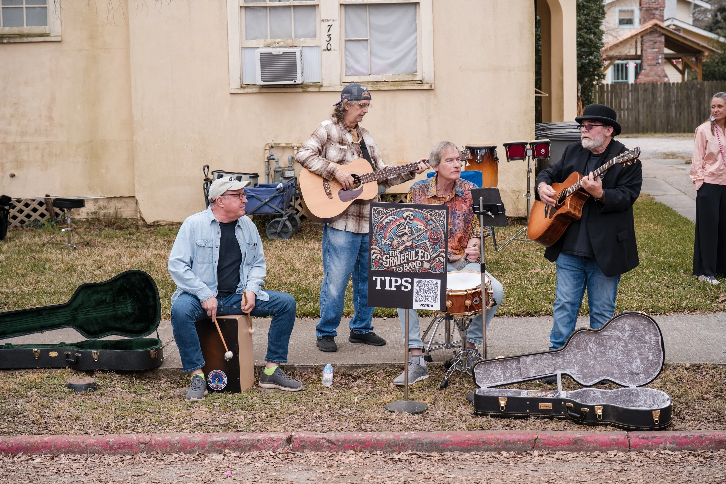 A group of five musicians performing on the sidewalk in front of a house. They are playing guitars, a cajón, and drums. There are open instrument cases on the ground. One person is sitting with drumsticks, while others are standing or sitting playing