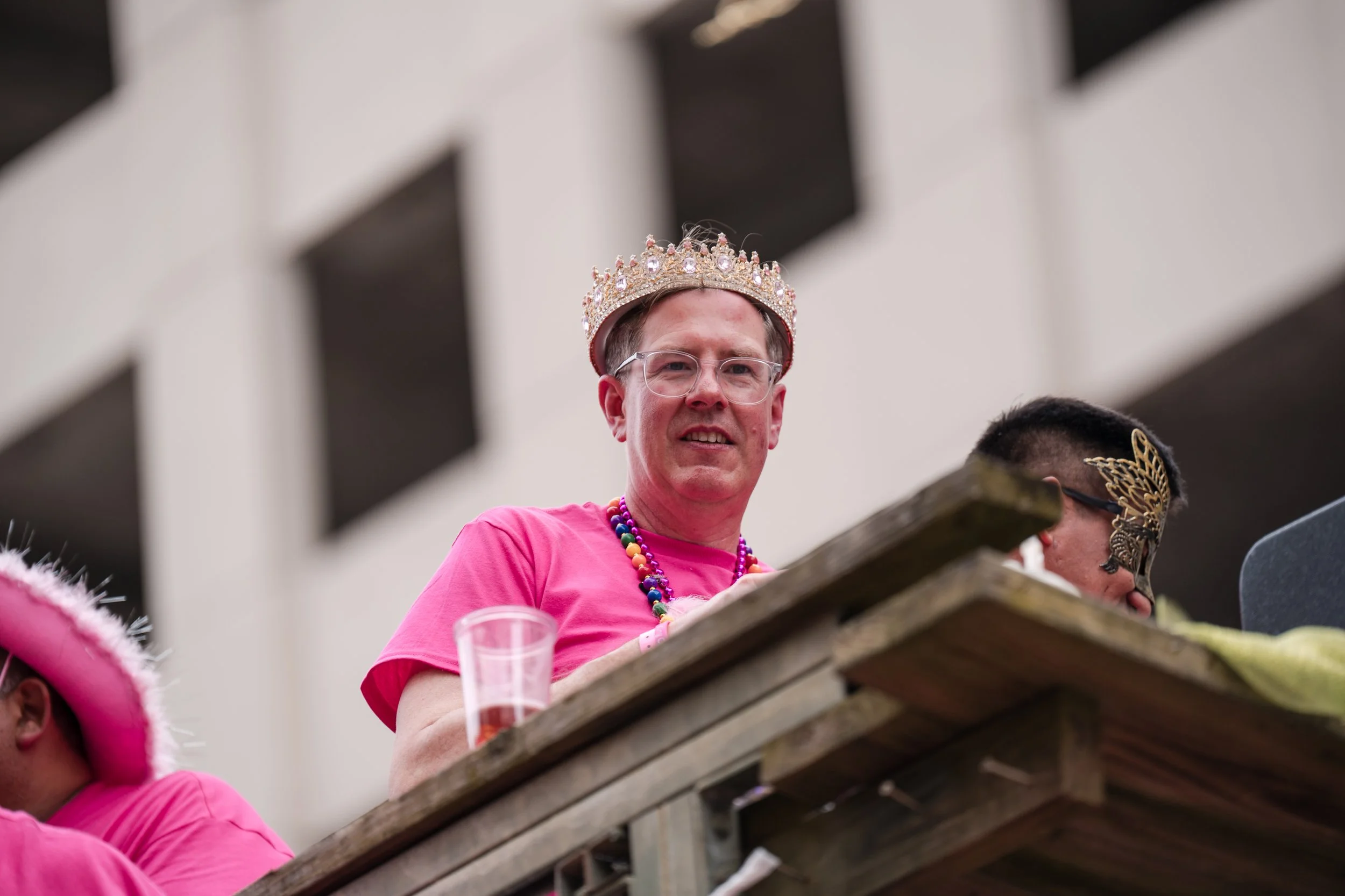 A man wearing a crown, glasses, and colorful beads is smiling at a parade or celebration. He is dressed in a pink shirt and is sitting on a float made of wood. There are other people around him, some dressed in pink and wearing decorative headwear.