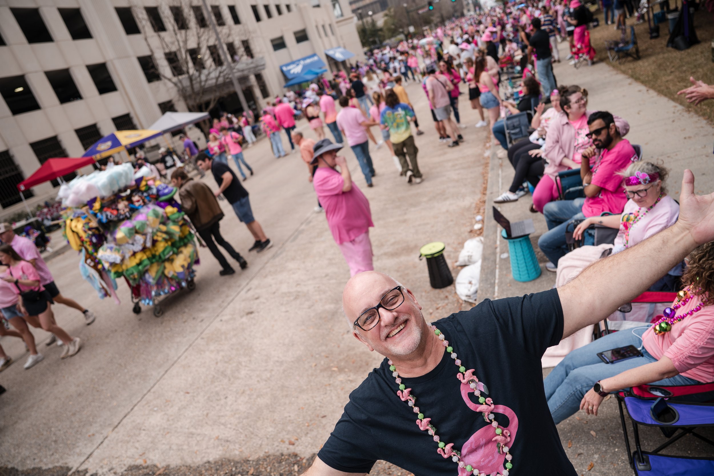 A man wearing glasses and a black shirt smiling and cheering at a crowded outdoor event. Many people are dressed in pink, celebrating in a city plaza with vendor tents and a large crowd in the background.