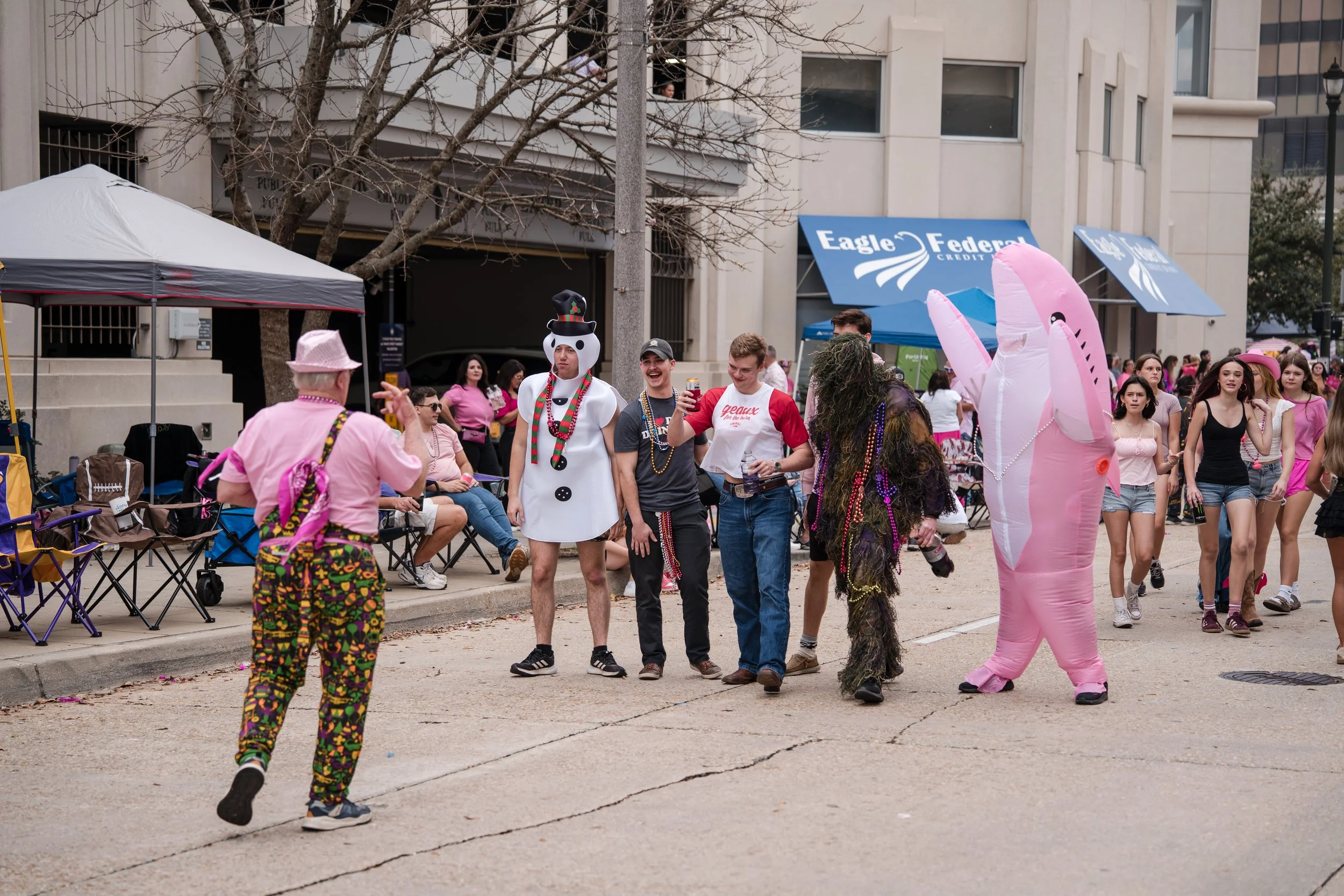 People dressed in costumes at a street event, including a snowman and a pink inflatable bunny, with onlookers sitting and standing along the sidewalk.