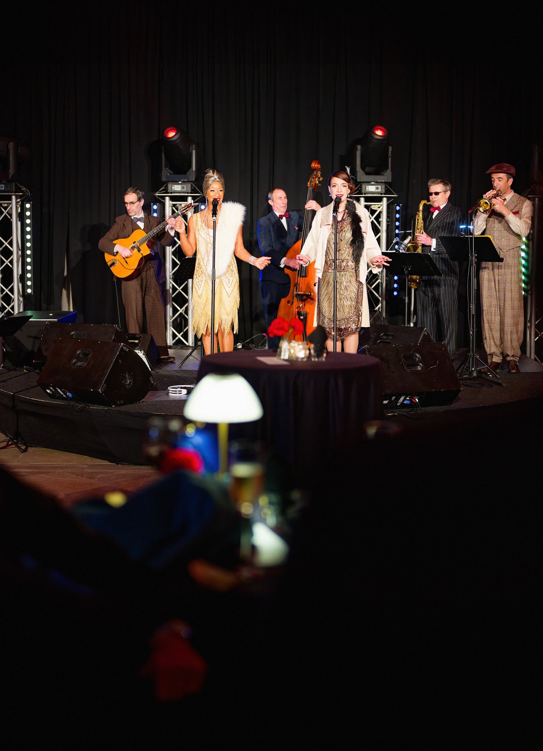 A live jazz band performing on stage with five musicians, two women singers, and three men playing guitar, double bass, saxophone, and trumpet, in a dimly lit venue with black curtains in the background.