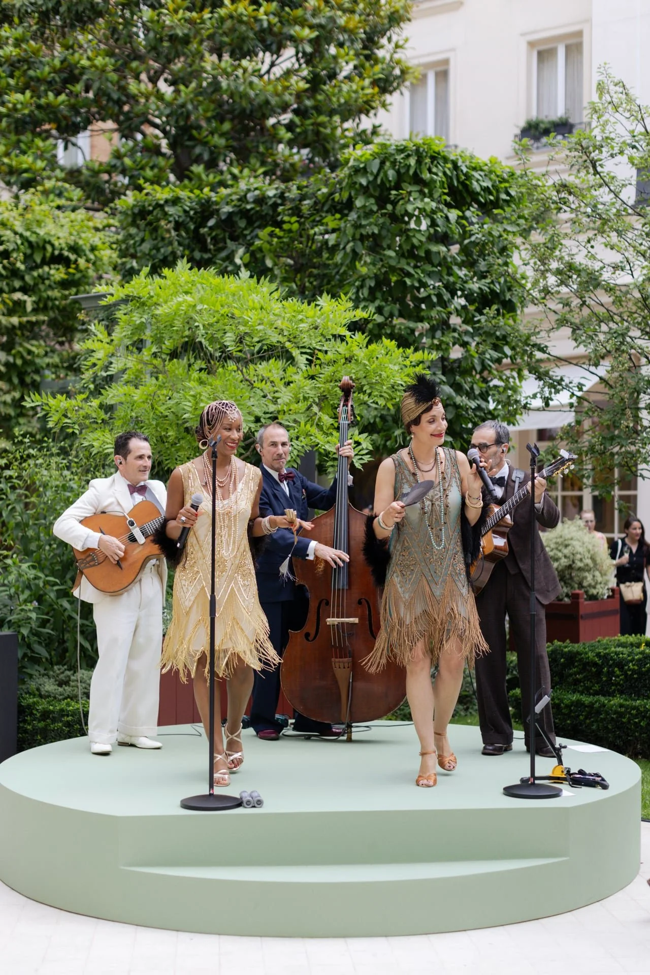 A vintage-style band performing on a small outdoor stage in a garden with lush greenery. The band includes a man in a white suit with a guitar, a woman in a gold flapper dress with a microphone, a man in a dark suit with a double bass, a woman in a fringed dress with a microphone, and a man in a dark suit with glasses playing an electric guitar.