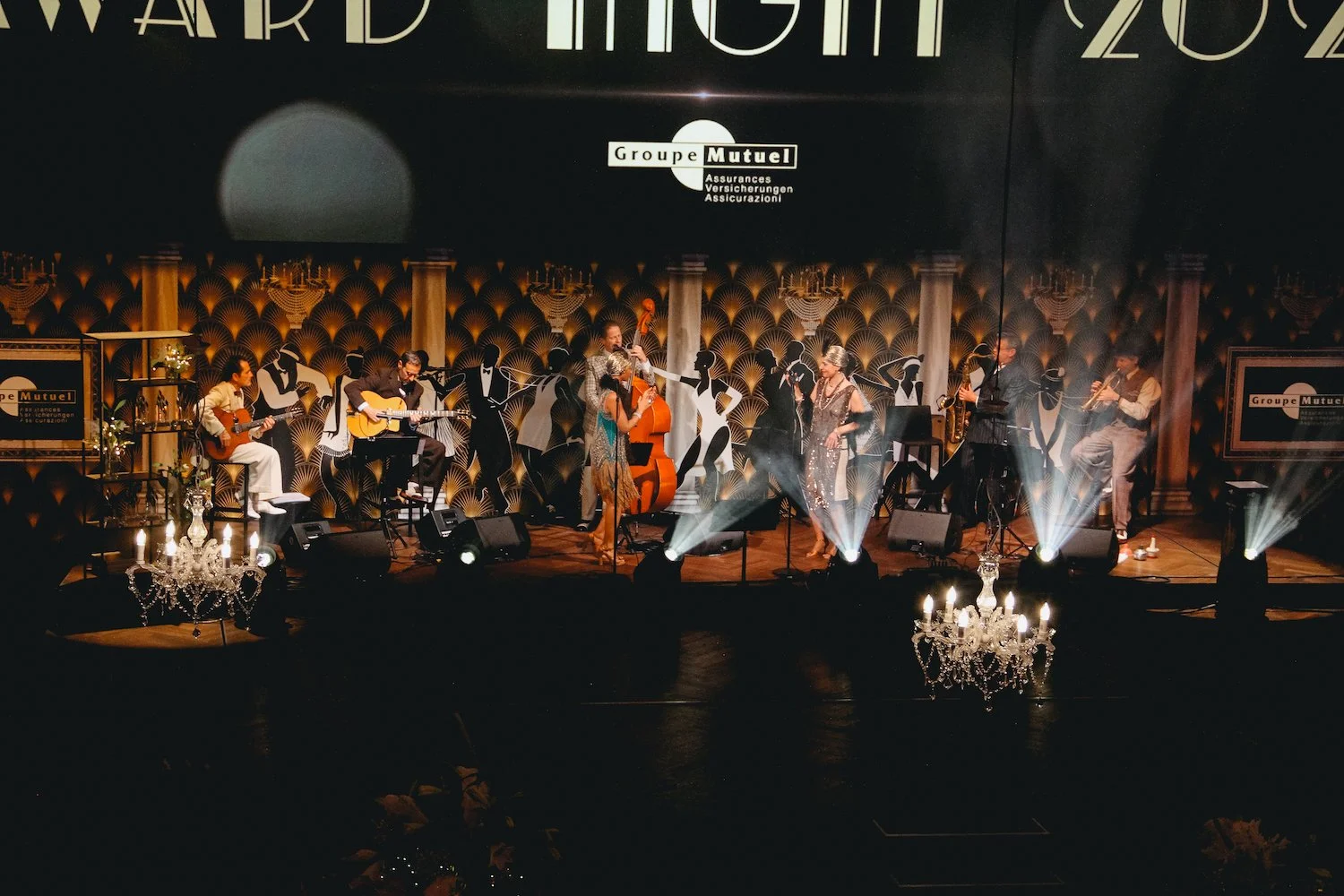 Stage with musicians performing in a ballroom, with elegant chandeliers, dark patterned wallpaper, and large signage for Grouper Mutual in the background.