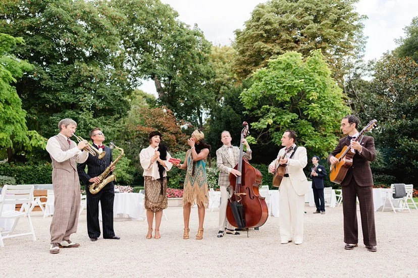A group of musicians playing instruments and singing outdoors in a garden setting for a celebration or event.
