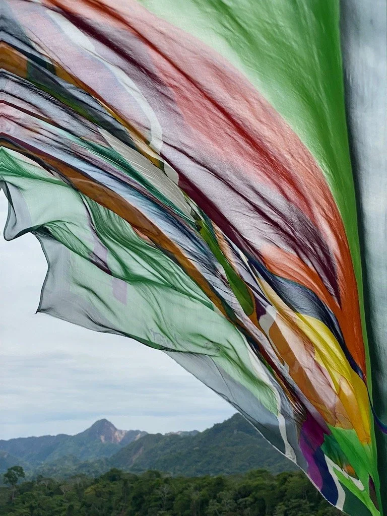 Colorful fabric fluttering in front of a mountain landscape with green hills and cloudy sky.