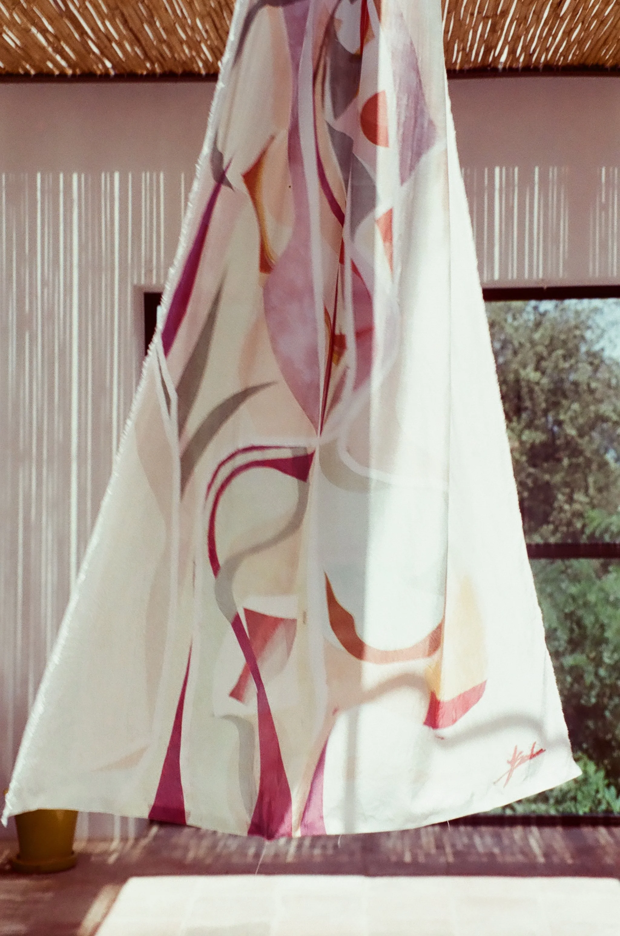 A fabric curtain with a floral or abstract design in pink, orange, and beige tones, hanging in front of a textured wall and window with trees outside.