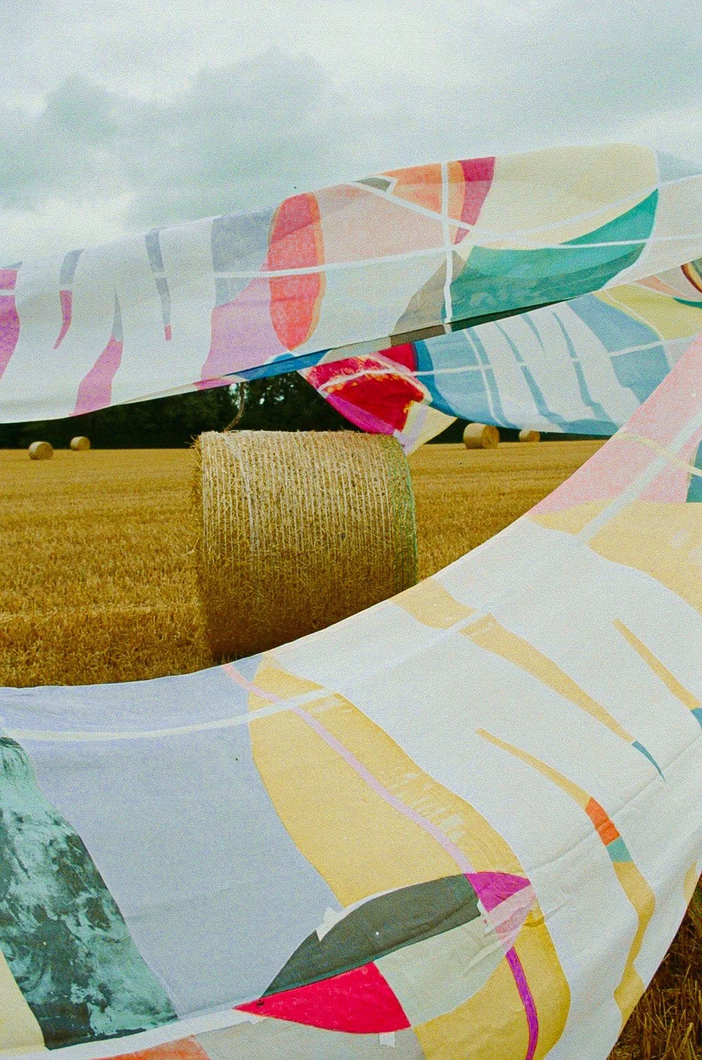 Colorful fabric with abstract patterns draped over hay bales in a field, with hay bales and a cloudy sky in the background.
