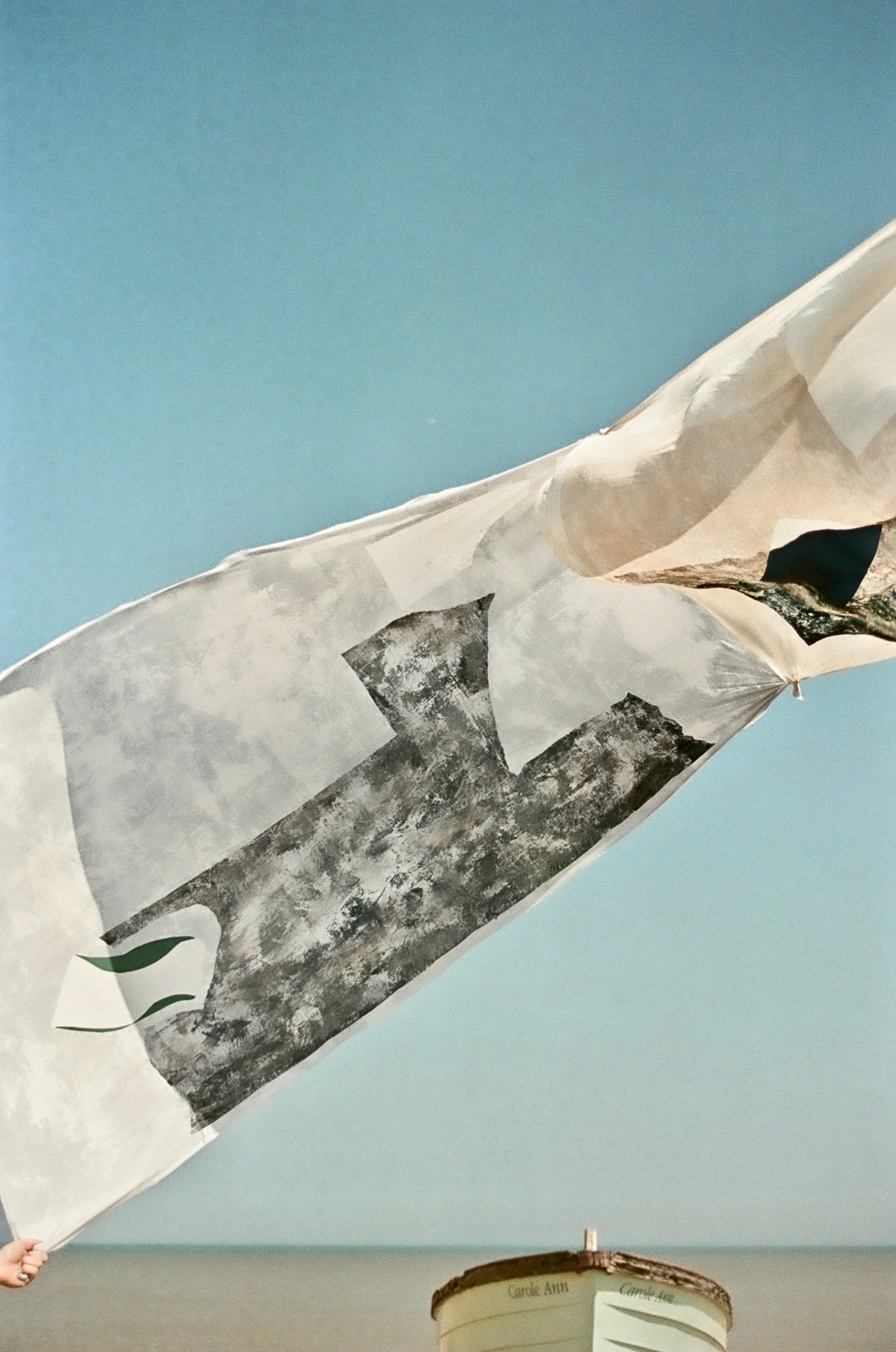 A large printed flag with a mountain landscape and sky, held by a person with a beaded bracelet, against a blue sky, with a small decorative trophy or bowl labeled 'Come Aim' on a surface below.