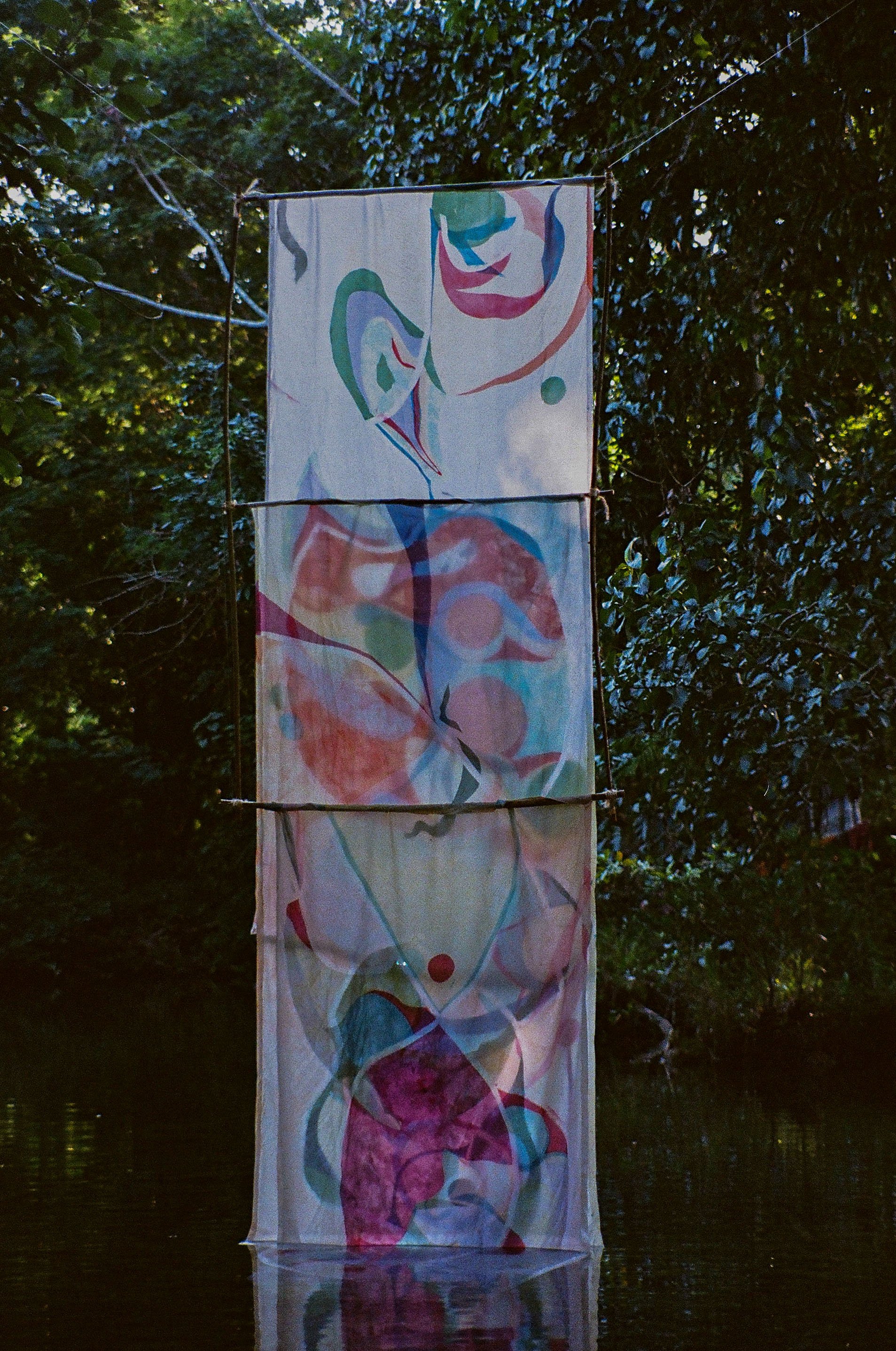A tall fabric banner with colorful abstract art is hanging outdoors near a body of water, surrounded by dense green trees.