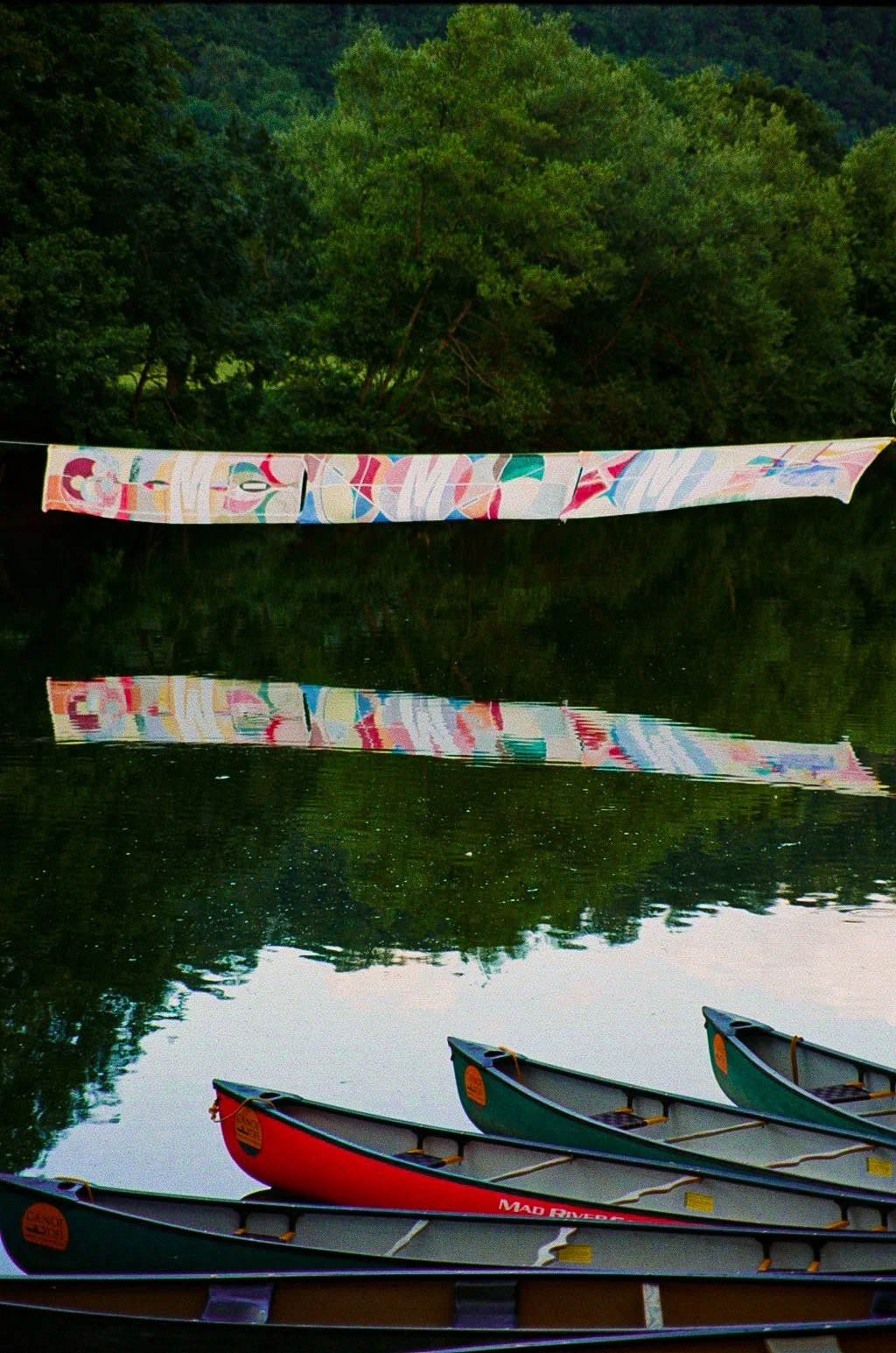Colorful tied-dye fabric hanging above calm river, with green trees in background and three canoes floating on the water.