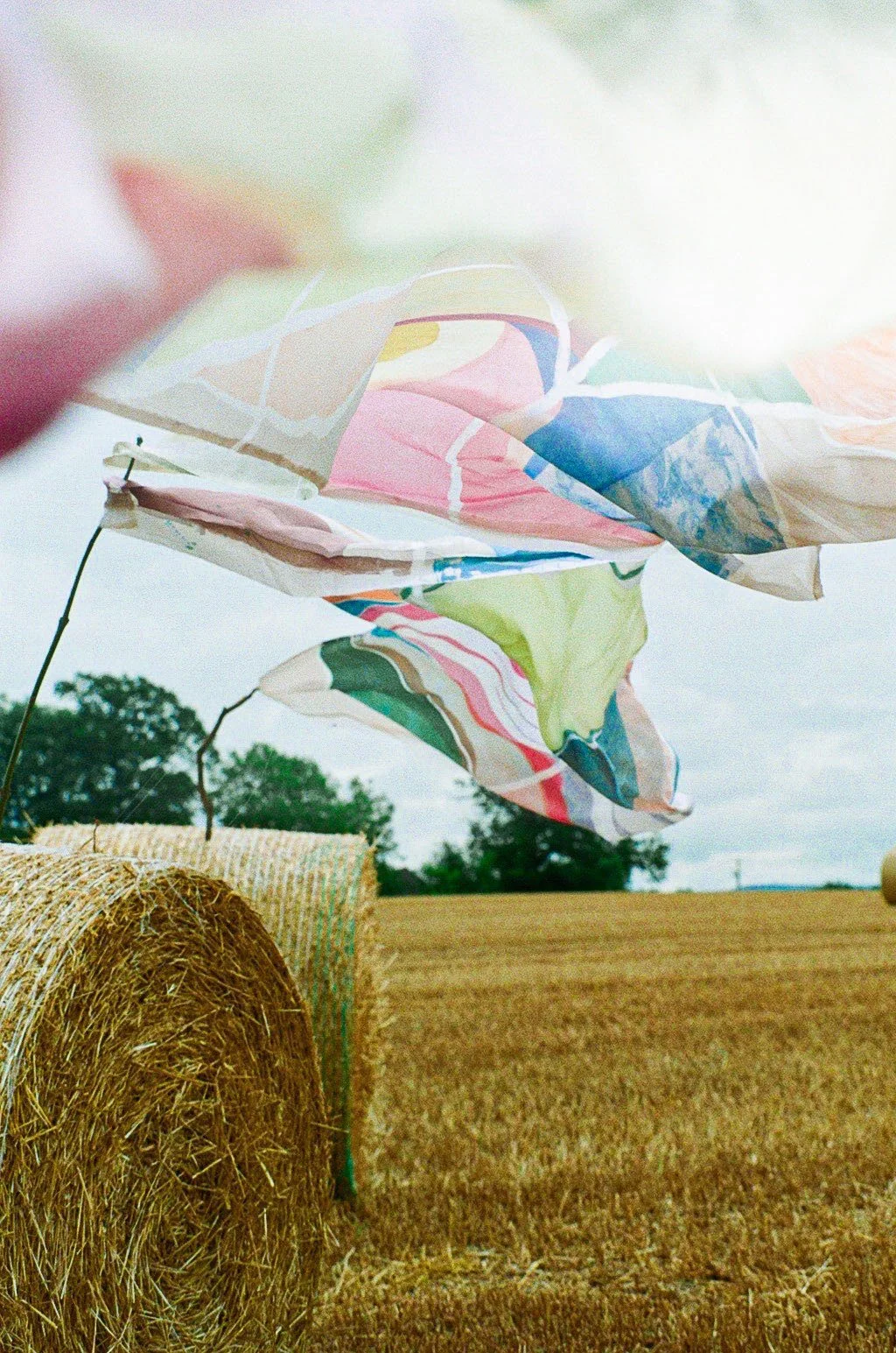 Colorful fabric or paper billowing in the wind above a hay bale in a rural field with overcast sky and trees in the background.