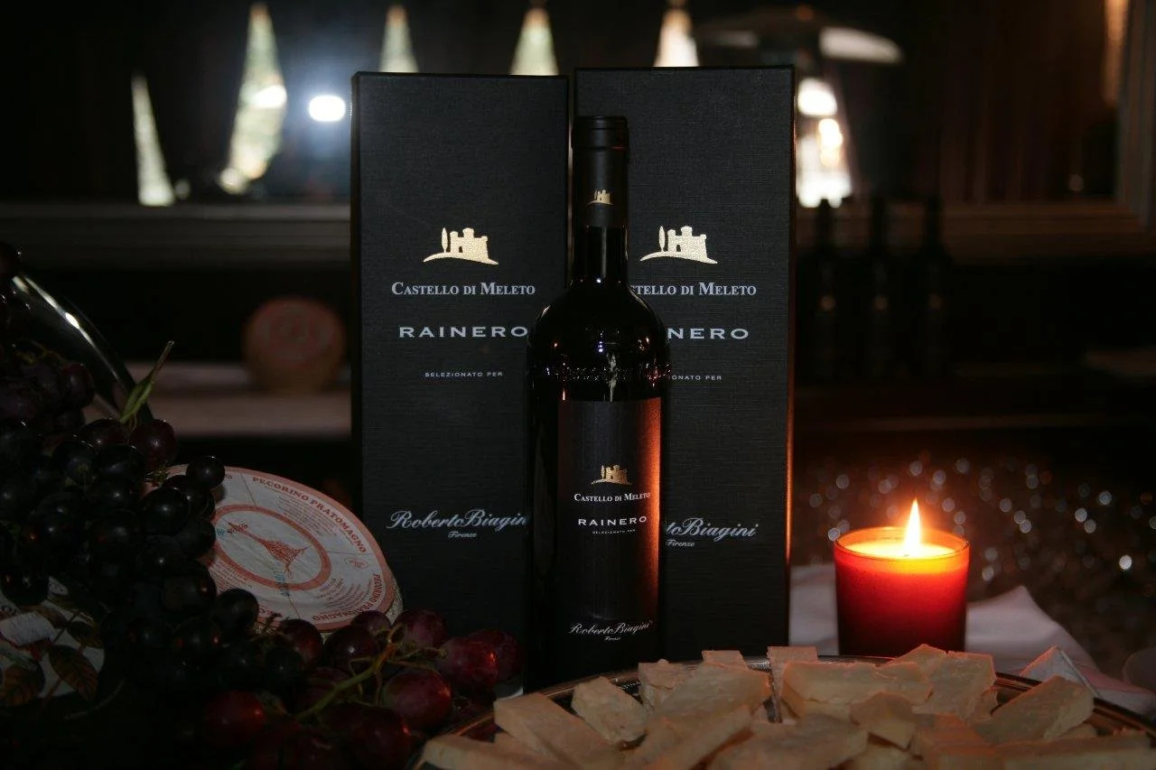 Bottiglia di vino con confezioni regalo sullo sfondo, grappoli di uva e formaggi circondano una candelaaccesa su un tavolo