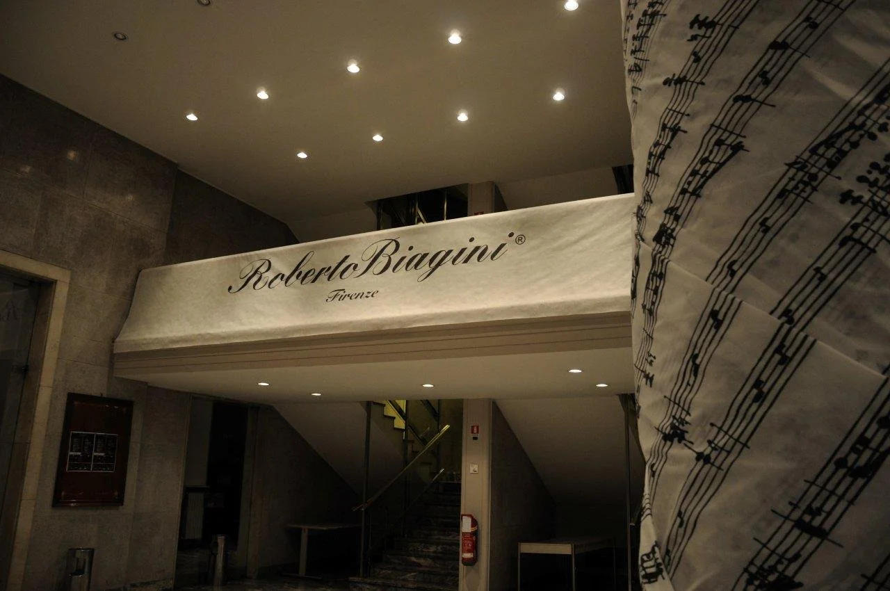 L'insegna di un negozio chiamato Roberto Biagini Firenze, con un soffitto illuminato e pareti di pietra, e una tuta con uno schema di note musicali in primo piano.