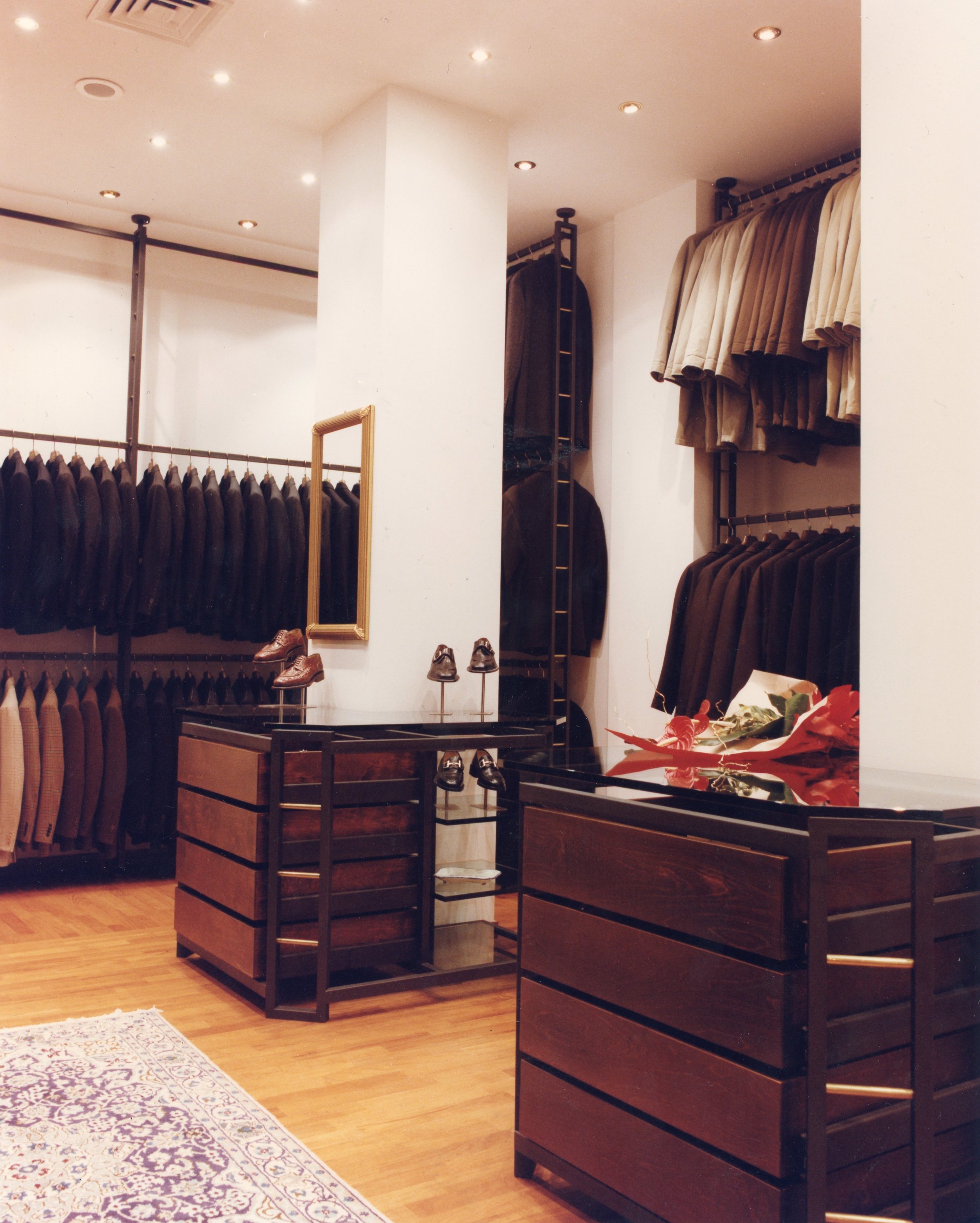 Interno di un negozio di abbigliamento con cappotti appesi, scarpe in esposizione, un grande specchio e un tappeto.