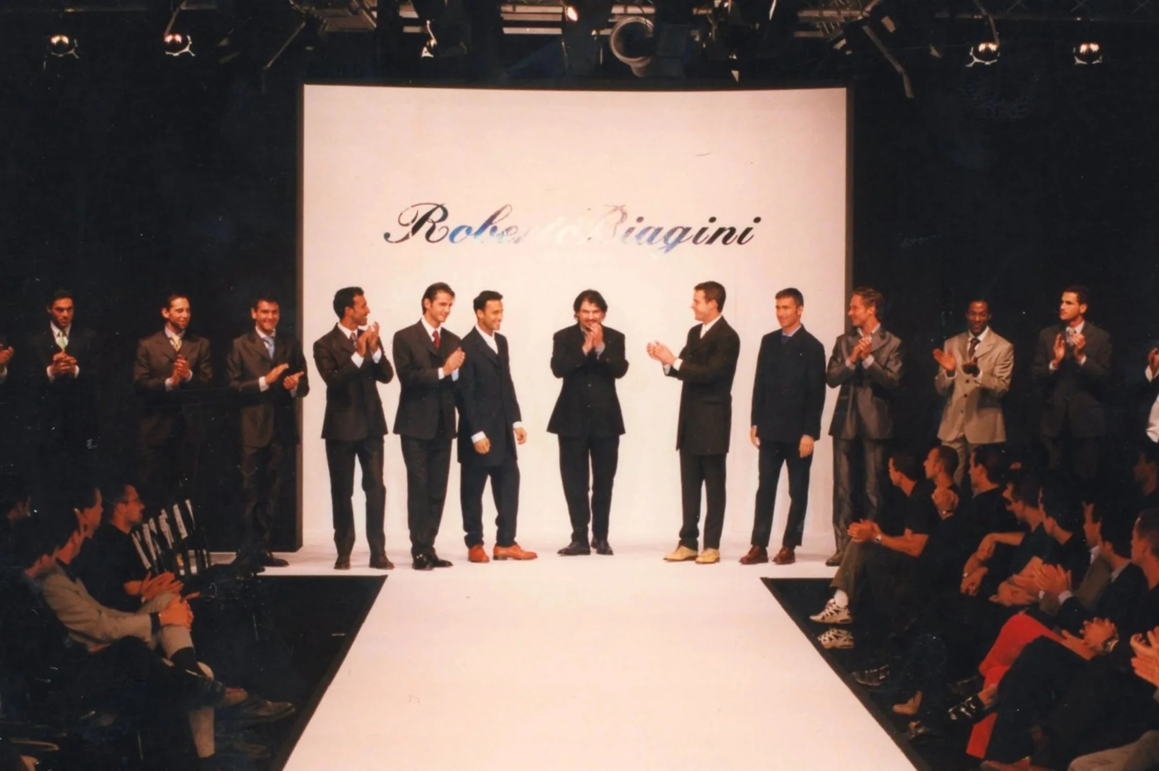 Un gruppo di modelli in abiti eleganti sul palco durante una sfilata di moda, con uno schermo in background che mostra la scritta 'Robe di Riagini', pubblico seduto ai lati.