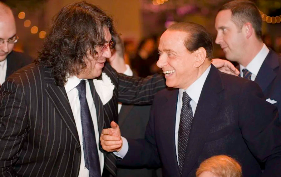 Due uomini in abiti eleganti si stringono la mano e sorridono durante un evento sociale, con altri presenti sullo sfondo.