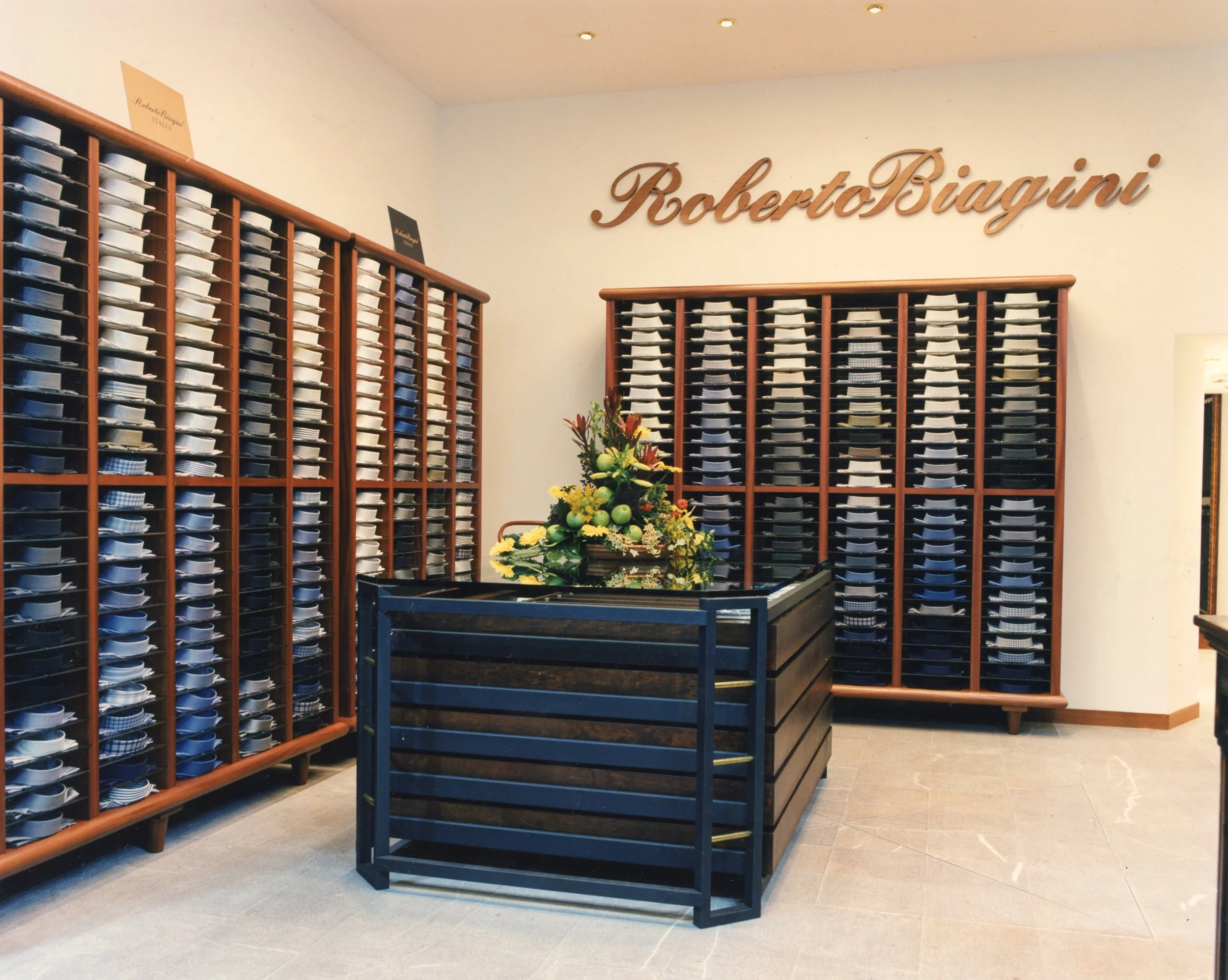 Ambiente di negozio di camicie con scaffali di piastrelle di camicie in vari colori e motivi, firma "Roberto Biagini" sulla parete e decorazione floreale al centro.