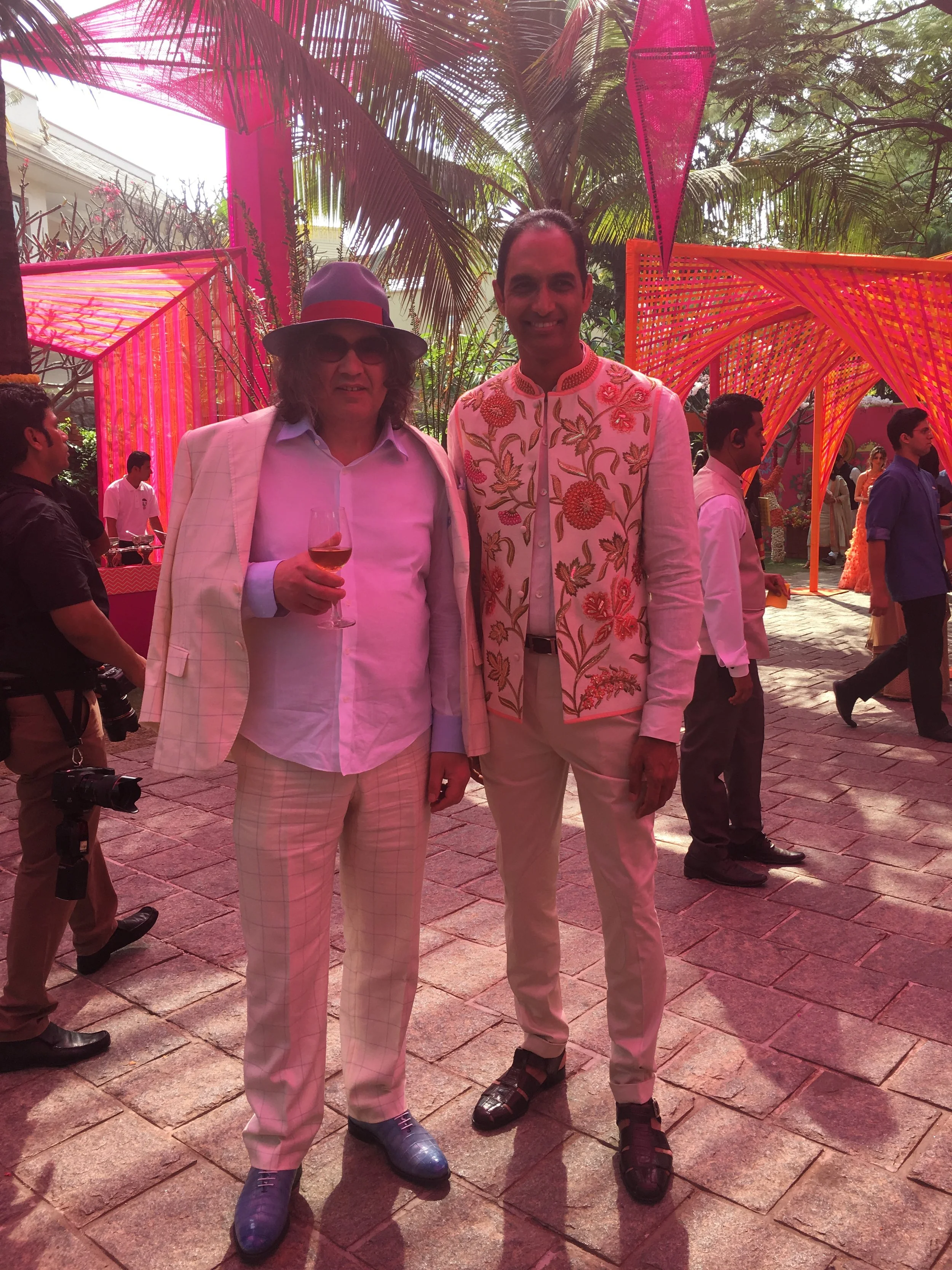 Due uomini indossano abiti eleganti e colorati in un evento all'aperto, con decorazioni di tessuto rosa e arancione e palme sullo sfondo. Uno dei partecipanti tiene un bicchiere di vino.