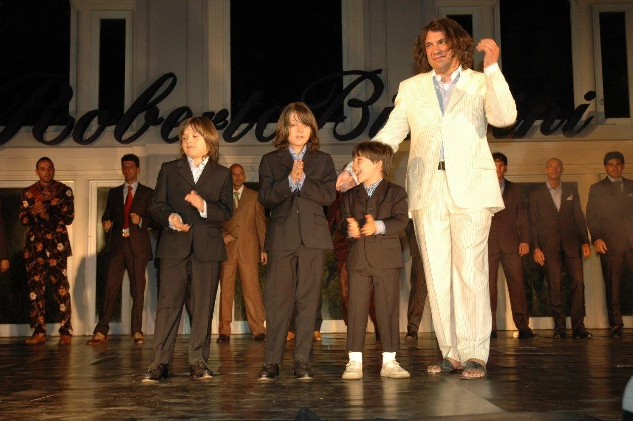 Sul palco, un uomo con capelli ricci e vestito a righe bianche e crema, accarezza tre bambini in abiti eleganti. Sullo sfondo, persone in abiti formali applaudono.