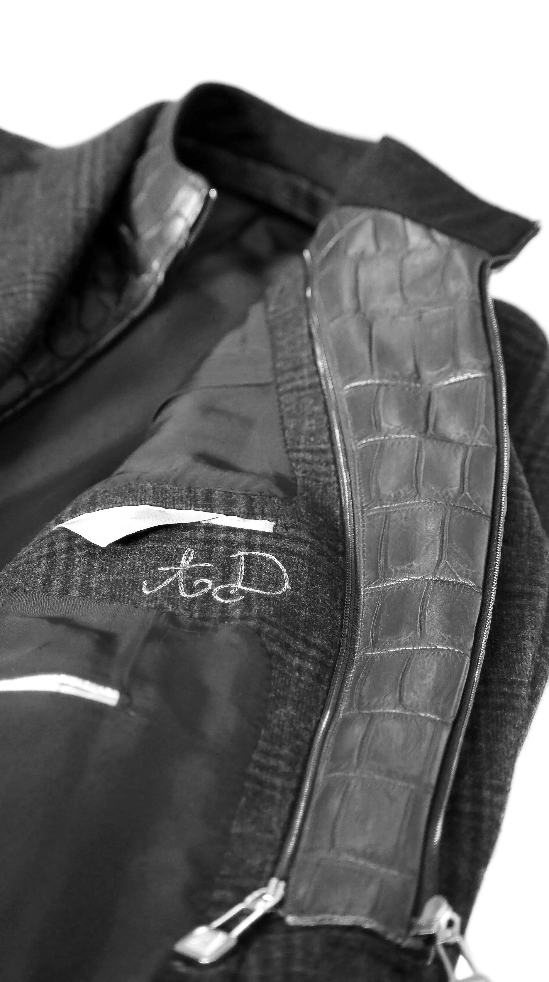 Primo piano di un cappotto di lana e di una giacca con dettagli in pelle con motivo coccodrillo, con una zip. Sono visibili anche alcune etichette e un linguetta con una scritta in corsivo.