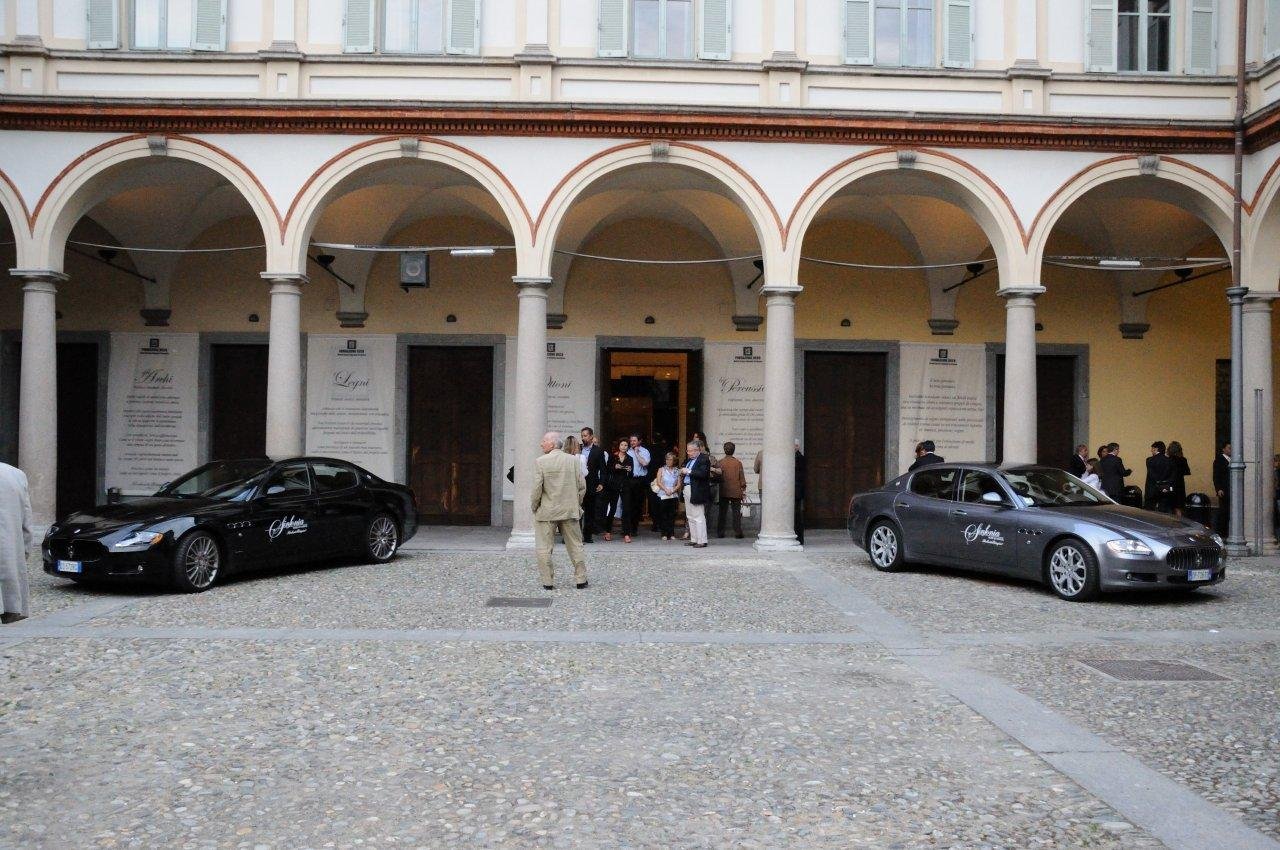 Vista del cortile di un edificio storico con arcate, due auto di lusso parcheggiate e alcune persone che parlano davanti all'ingresso.