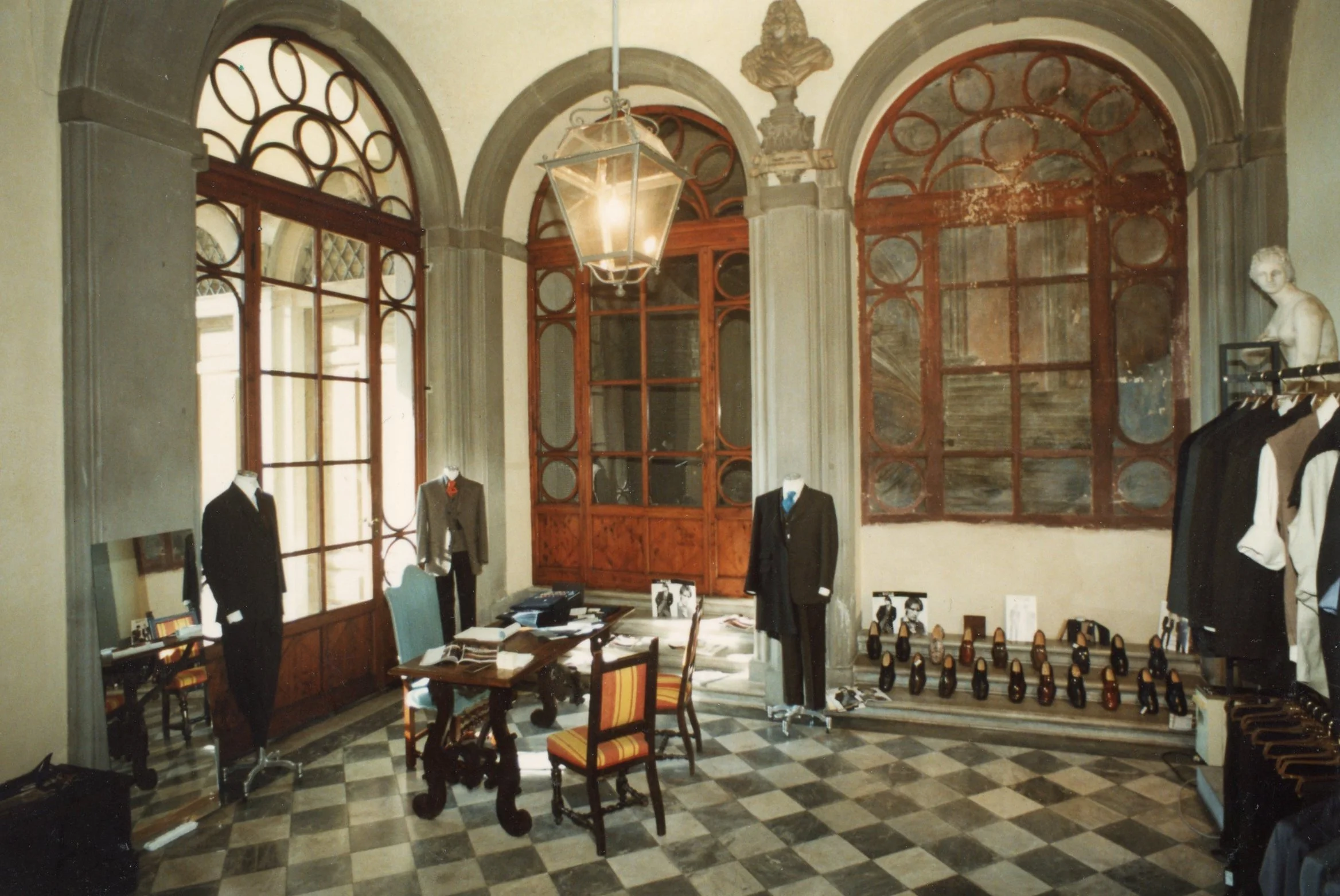 Interno di un negozio di abbigliamento con manichini vestiti eleganti, finestre grandi con struttura in vetro, lampadario di metallo, pavimento a scacchi, statue e scaffali con scarpe in fondo alla stanza.