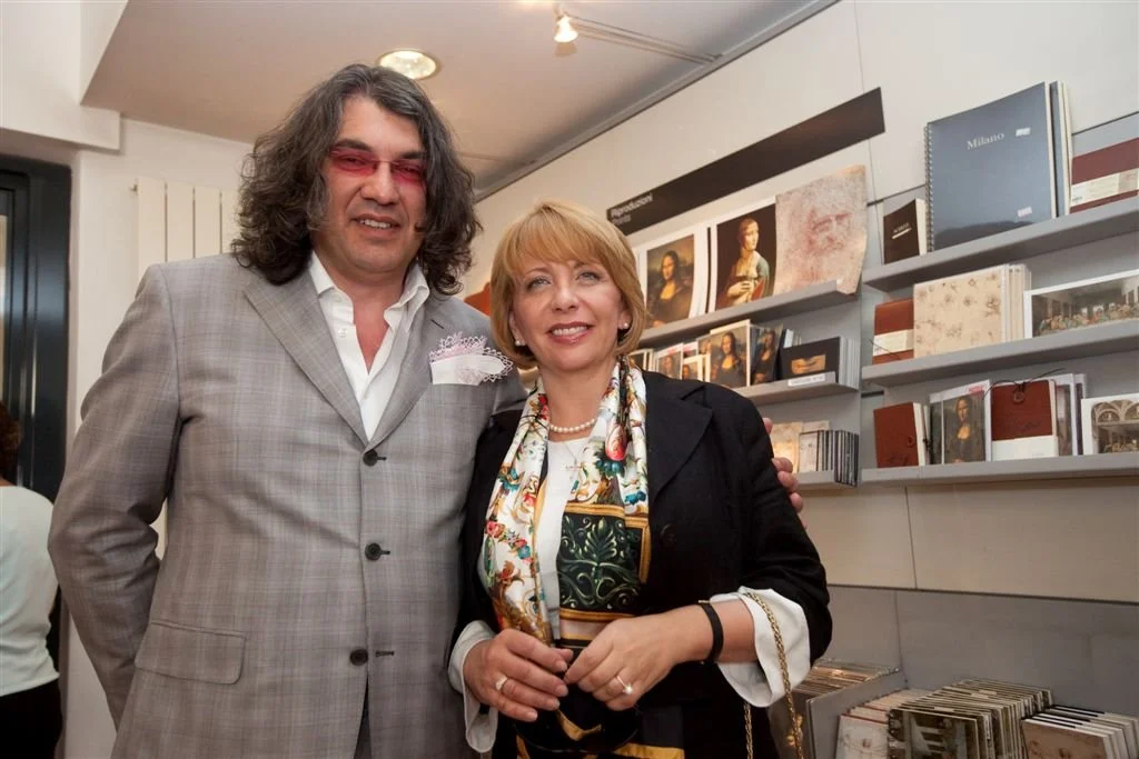 Un uomo con capelli lunghi e abito grigio, con gli occhi coperti da occhiali rossi, e una donna con cappotto nero e sciarpa colorata in un'anteprima di mostra di libri e fotografie in un'illuminazione artificiale in un ambiente artistico o culturale.
