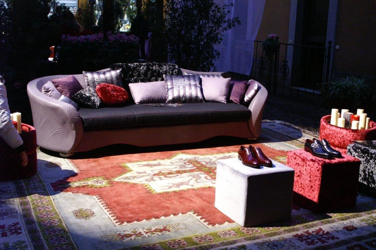 Soggiorno elegante con divano colorato, cuscini di varie forme e colori, tappeto decorativo, ciottoli e scarpe in primo piano, e candele in un contenitore rotondo rosso.