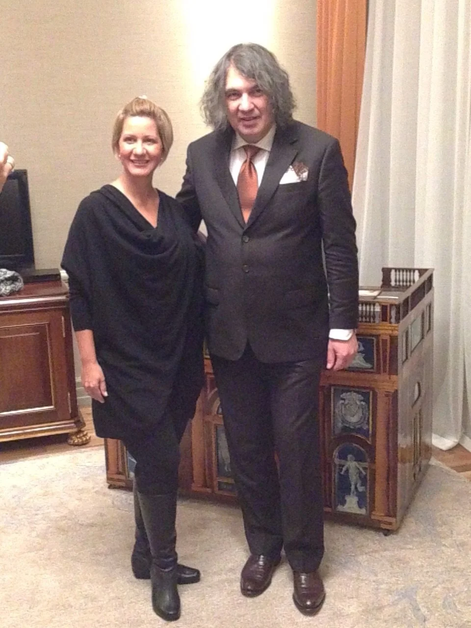 Una donna e un uomo in abiti eleganti si incontrano sorridendo in una stanza, con mobili di legno alle spalle e una tenda bianca a destra.