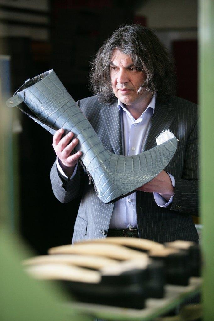 Uomo con capelli ricci e grigi che indossa un completo a righe, guarda un oggetto che assomiglia a una scarpa o calzatura di eleganza, con uno sfondo sfocato.