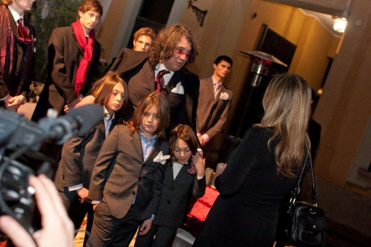Un gruppo di persone vestite elegantemente, in un ambiente interno, sembrano partecipare a un evento o a una cerimonia. Una donna con i capelli lunghi e biondi si trova di schiena, mentre alcuni bambini e un uomo con capelli ricci sono rivolti verso 