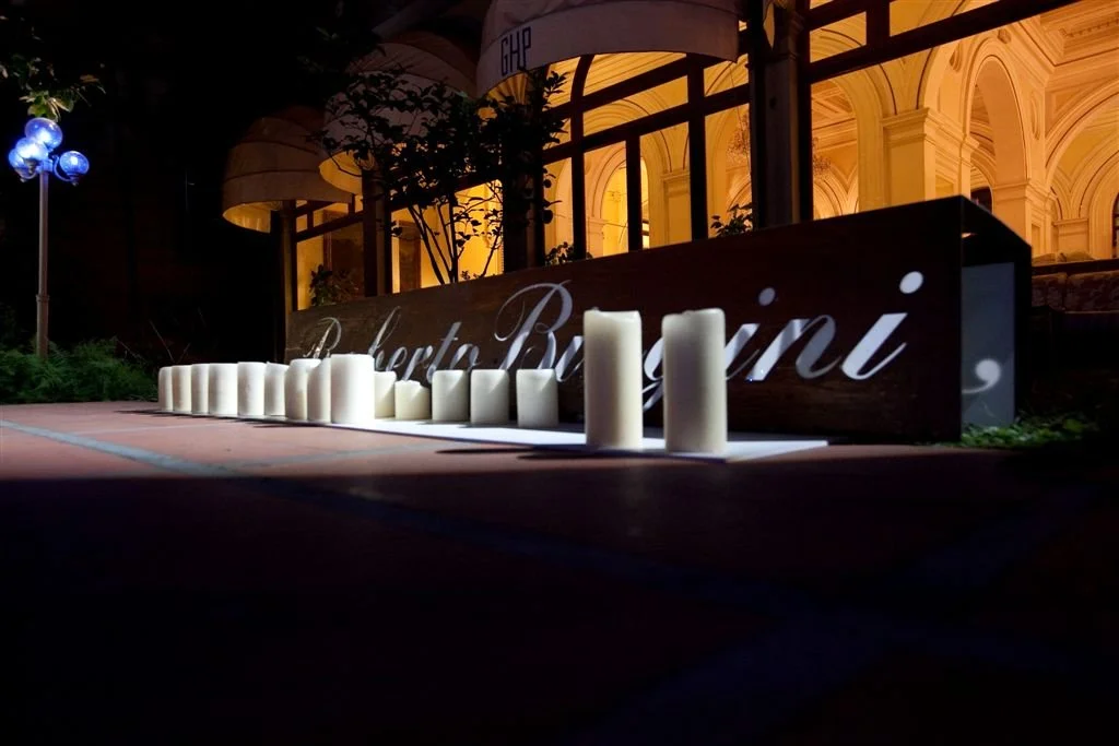 Segnaletica luminosa con candele bianche in una piazza di notte, sullo sfondo un edificio con archi e luci calde