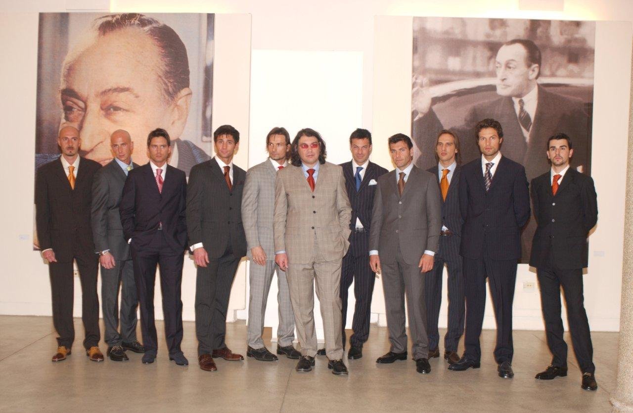 Un gruppo di uomini eleganti in abiti da sera, alcuni con cravatte, posano in fila in un museo o galleria d'arte, con grandi fotografie di uomini famosi sullo sfondo.