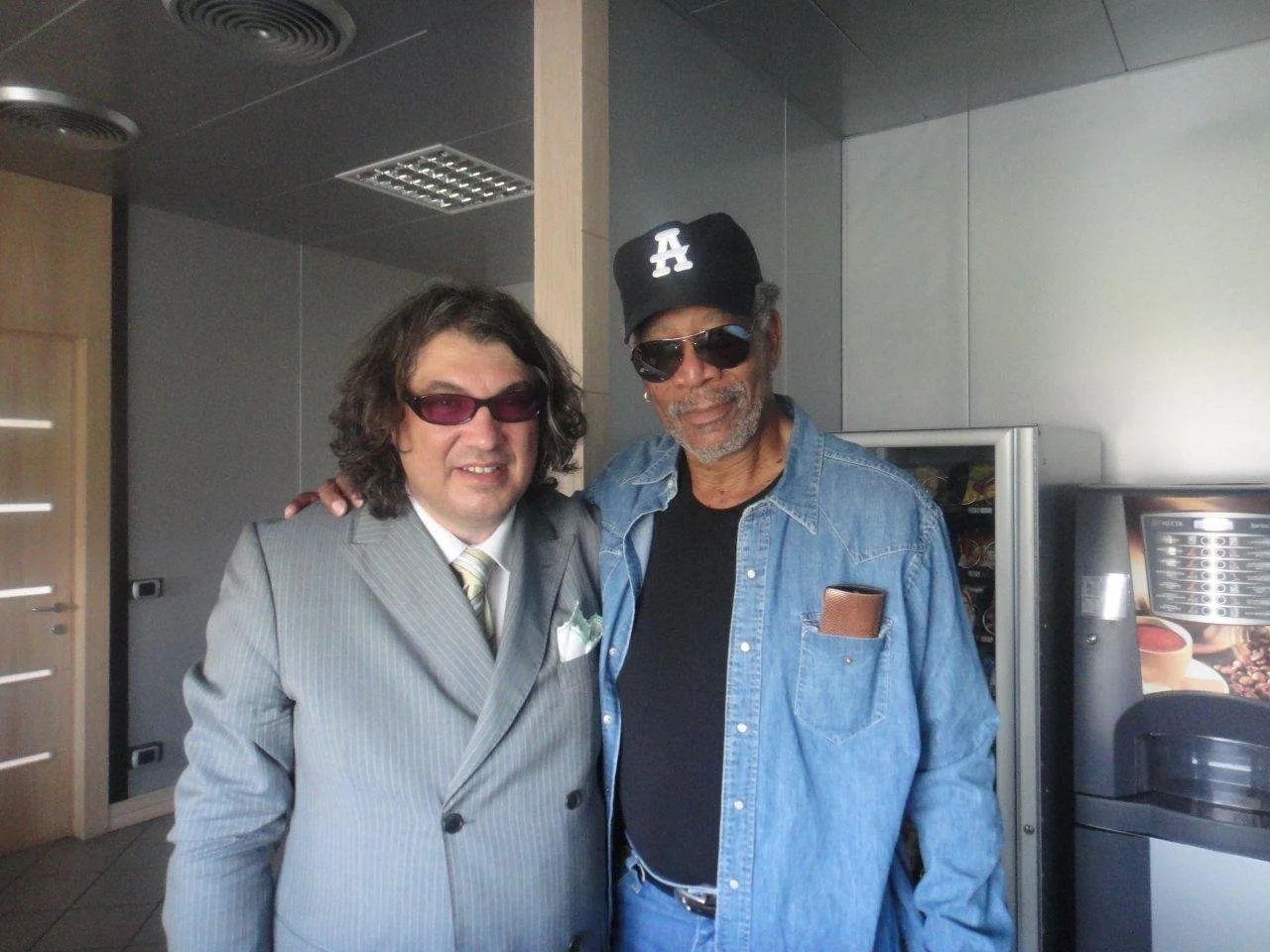 Due uomini sorridenti indossano occhiali da sole e si abbracciano in un ambiente interno, uno in giacca grigia e l'altro in camicia di jeans e cappellino nero.