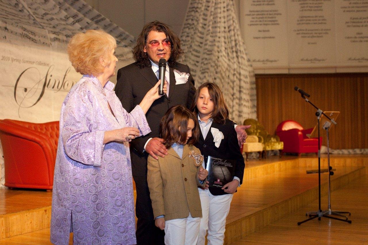 Una donna anziana che tiene un microfono e parla con un uomo vestito elegantemente, accompagnato da due bambini, uno dei quali tiene un premio. Sono su un palco in un ambiente di evento o cerimonia.