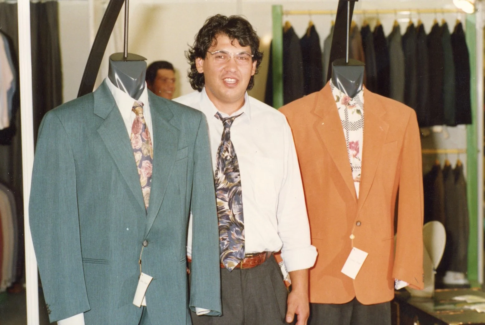 Uomo con capelli ricci e occhiali che si trova tra due manichini con abiti eleganti, in un negozio di abbigliamento.